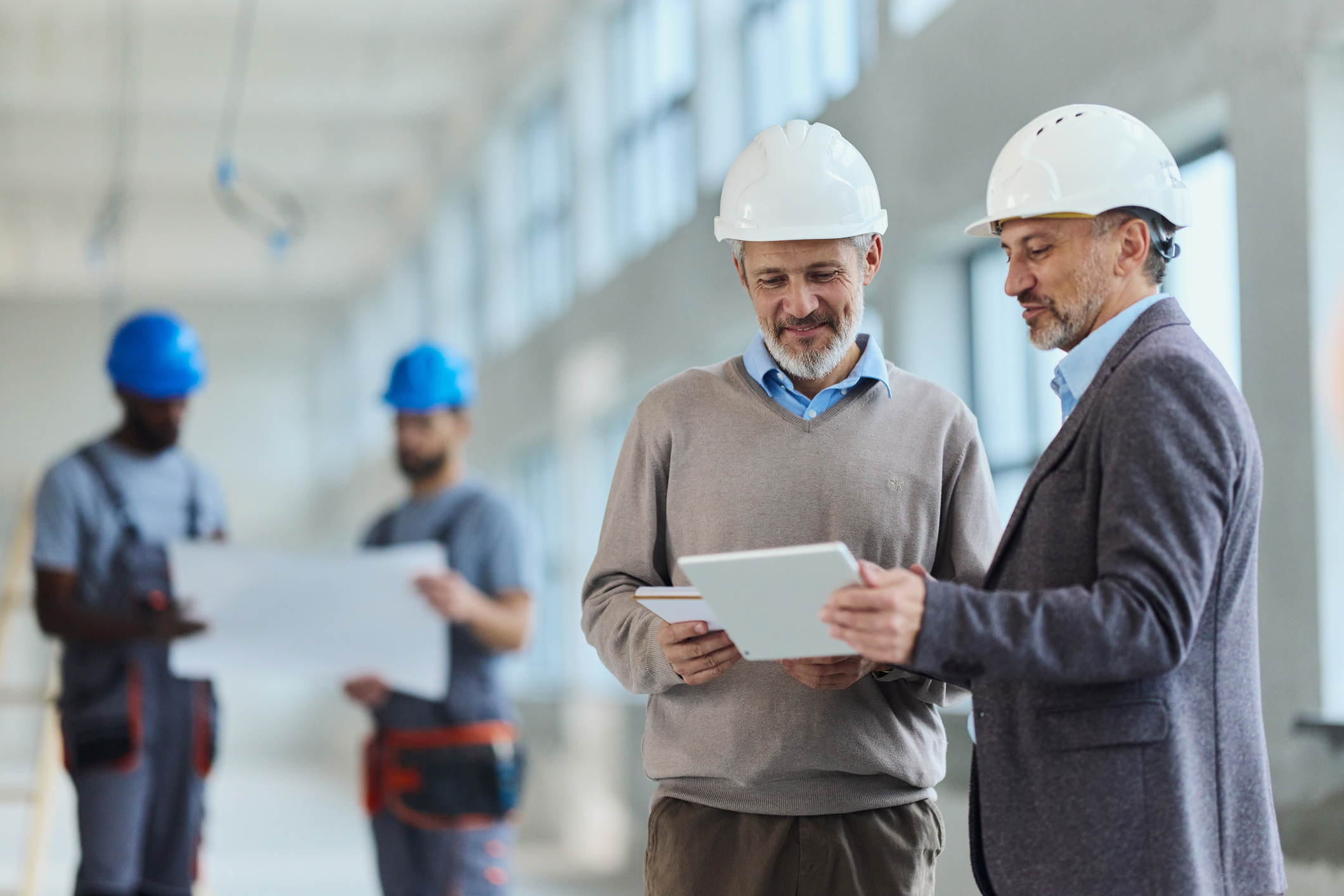 Two men in suits and white hard hats review documents on a tablet at a construction site, while two workers in blue helmets examine blueprints in the background.