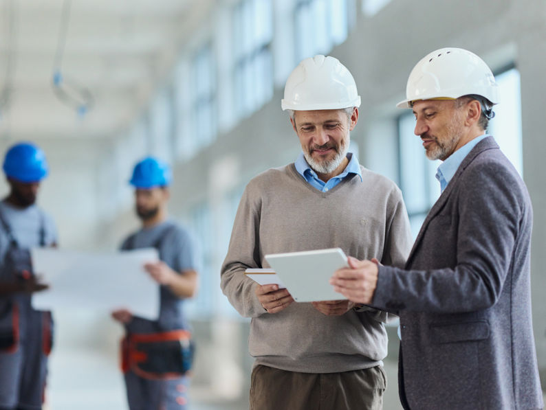 Two men in suits and white hard hats review documents on a tablet at a construction site, while two workers in blue helmets examine blueprints in the background.