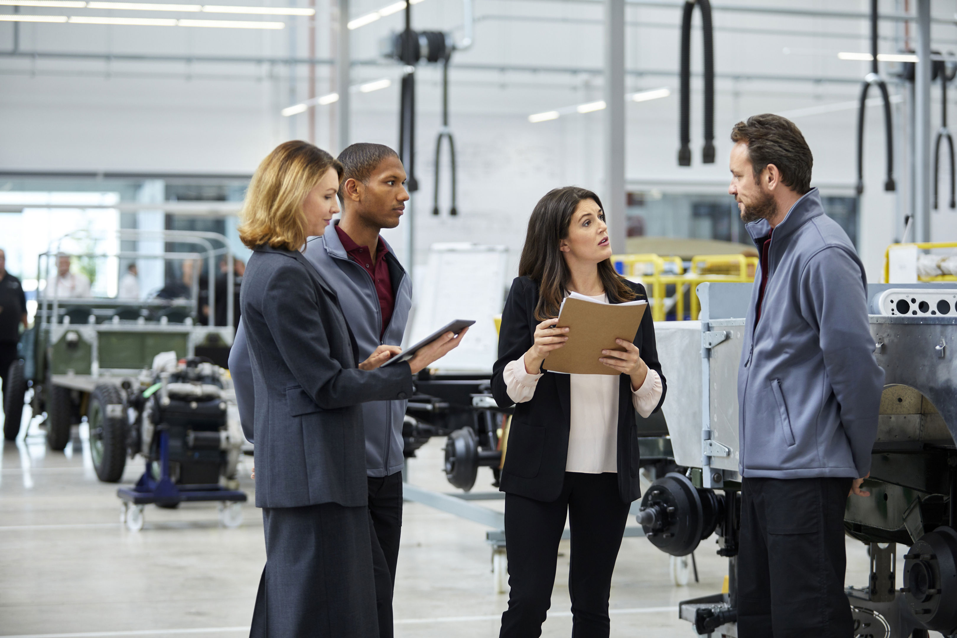 Four professionals stand in a modern factory, engaged in discussion. Two hold clipboards, one uses a tablet, and they appear to be reviewing plans or data among industrial equipment and vehicle frames.