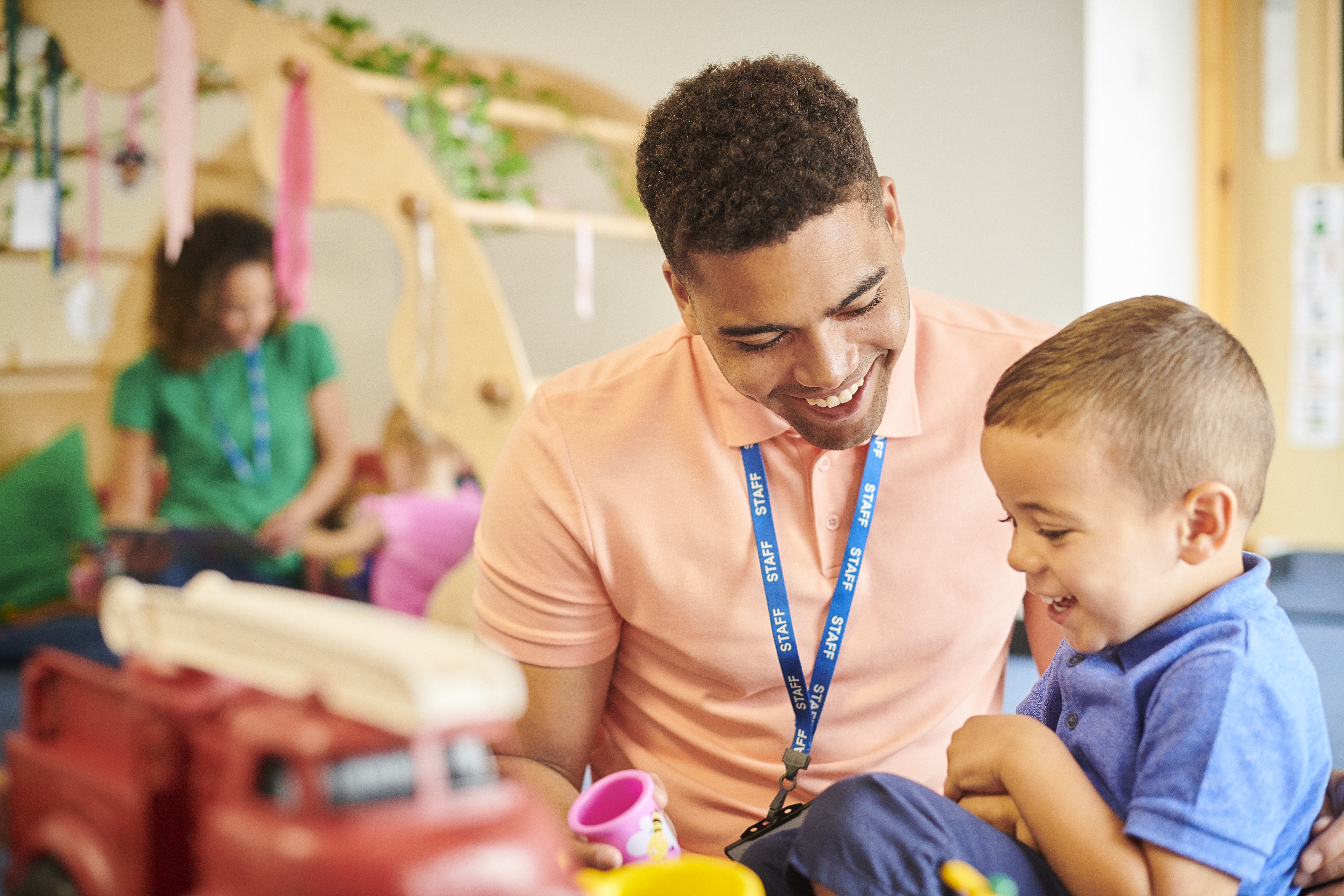 A smiling adult in a pink shirt plays with a cheerful young boy at a table with toys, including a red fire truck, in a bright, welcoming classroom setting.