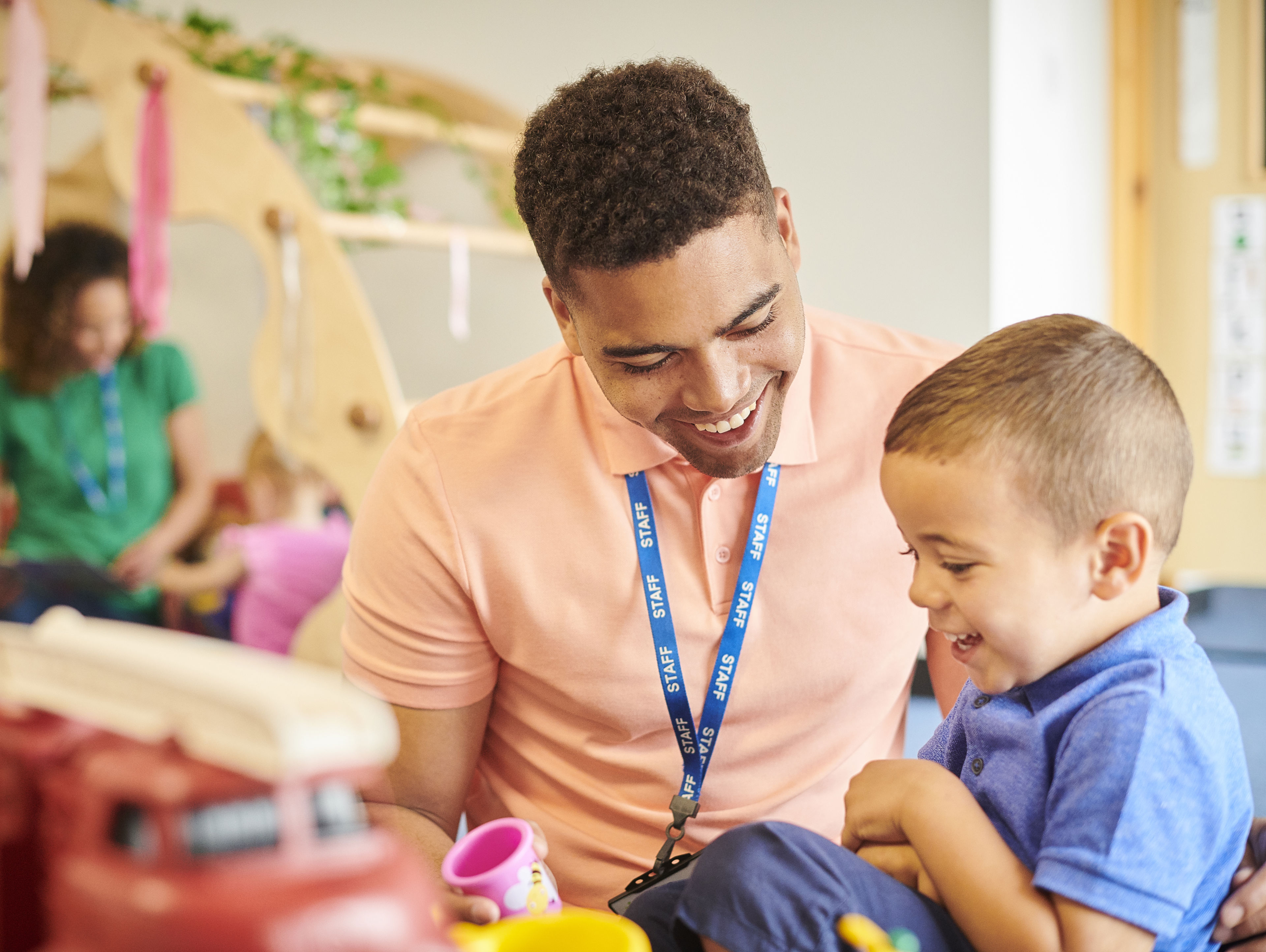 A smiling adult in a pink shirt plays with a cheerful young boy at a table with toys, including a red fire truck, in a bright, welcoming classroom setting.