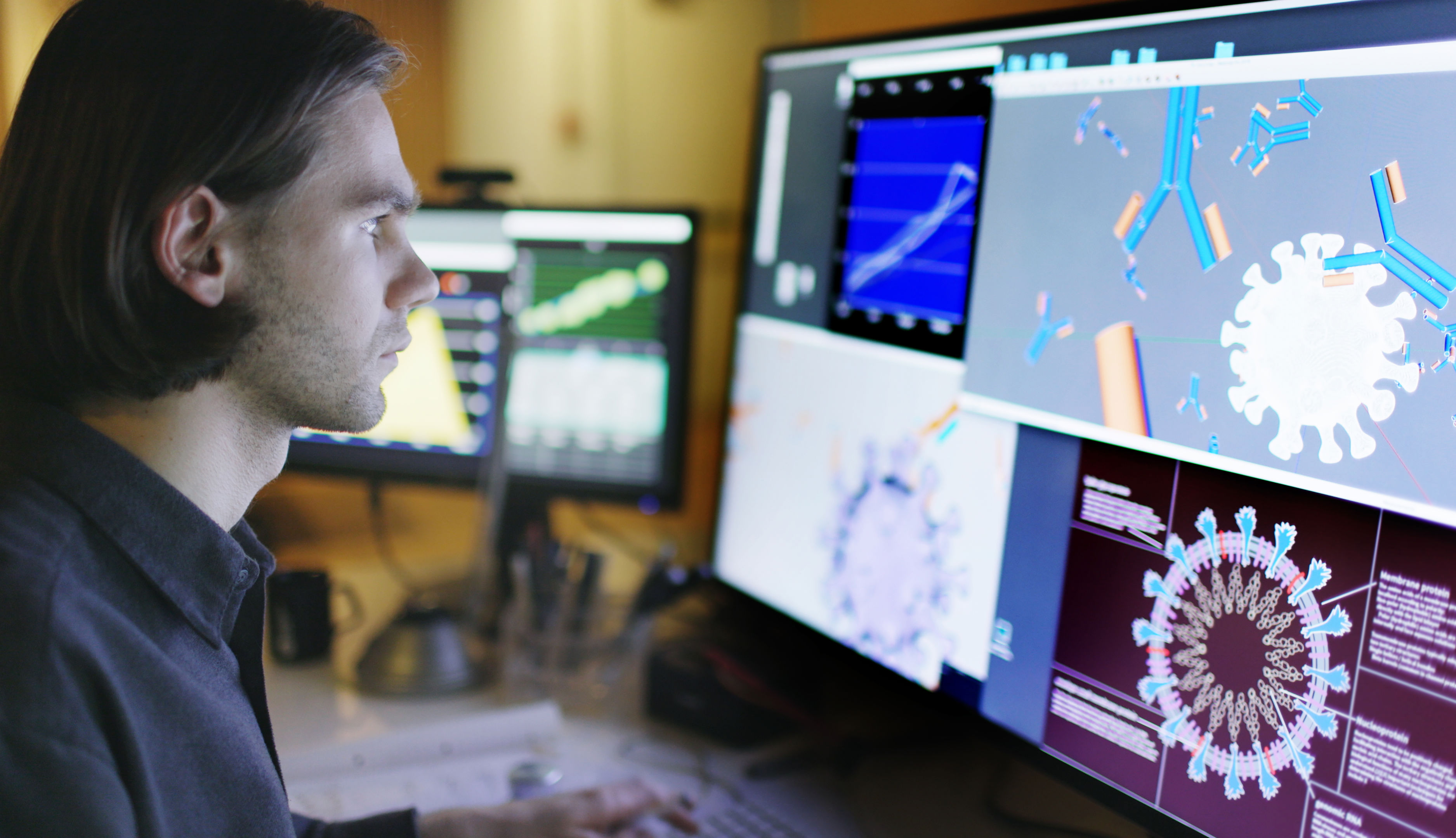 A person sits at a desk looking at computer monitors displaying colorful scientific diagrams and models of viruses and data charts, suggesting research or analysis in a laboratory or office setting.