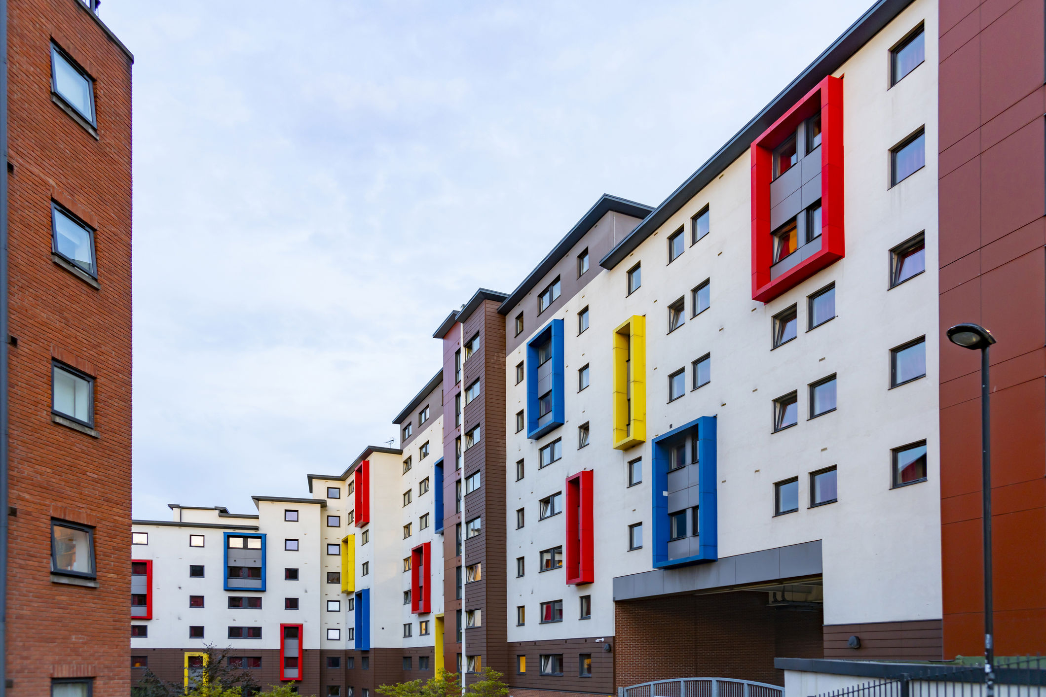 A modern apartment complex with white walls and colorful rectangular accents in red, blue, and yellow. The buildings have multiple stories and are connected by walkways. A clear sky is visible above.