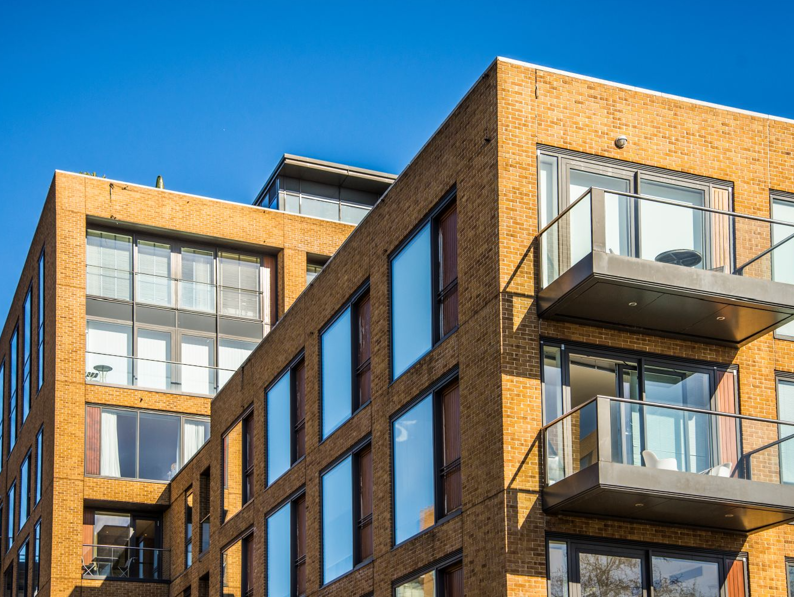 Modern apartment building with large windows and glass balconies, featuring a yellow-brown brick exterior under a clear blue sky.