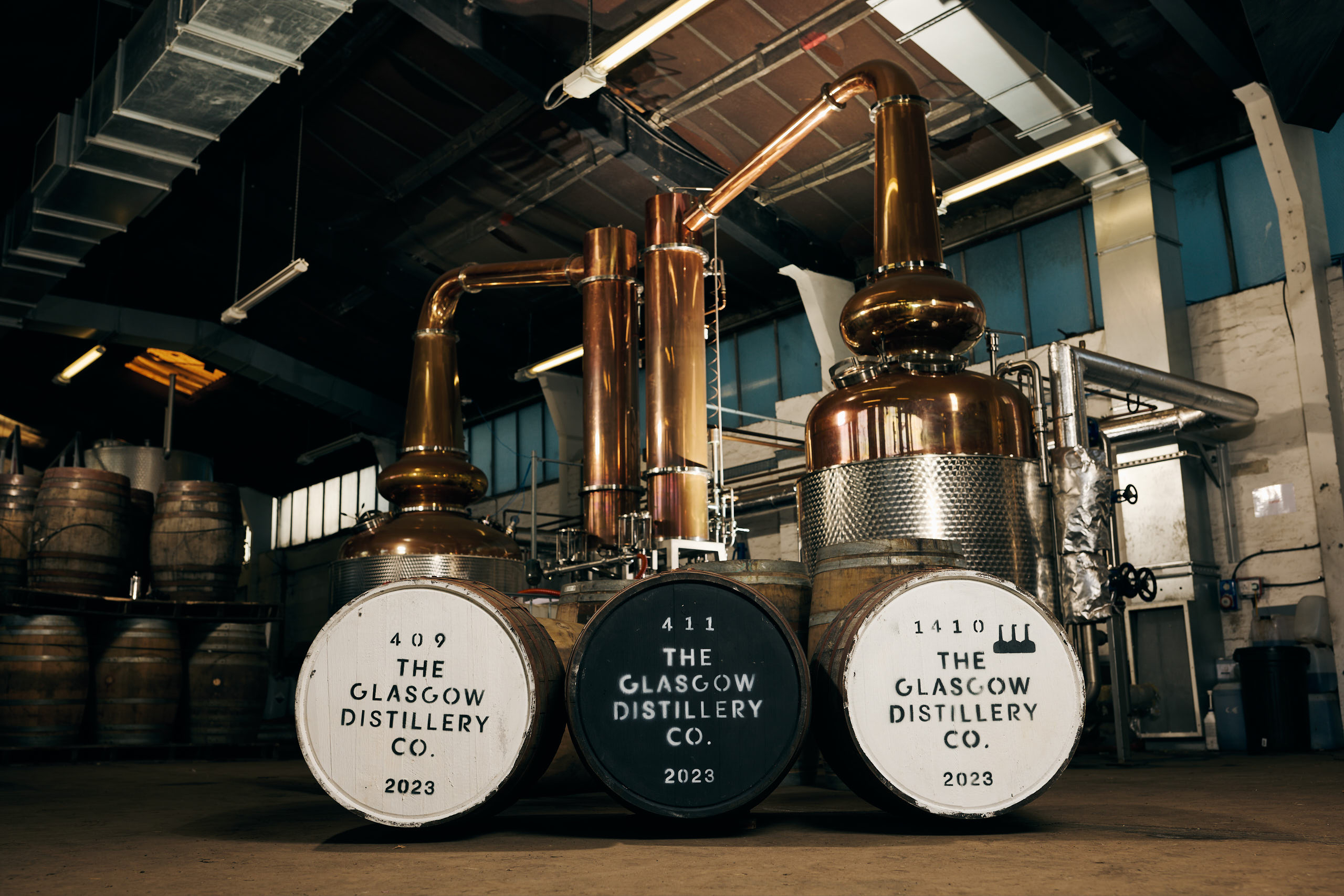 Copper stills and three large whisky barrels labeled “The Glasgow Distillery Co. 2023” are inside an industrial distillery with wooden casks stacked in the background and sunlight streaming from windows above.
