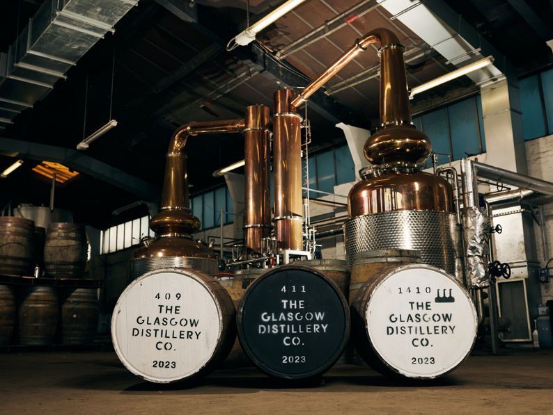 Copper stills and three large whisky barrels labeled “The Glasgow Distillery Co. 2023” are inside an industrial distillery with wooden casks stacked in the background and sunlight streaming from windows above.