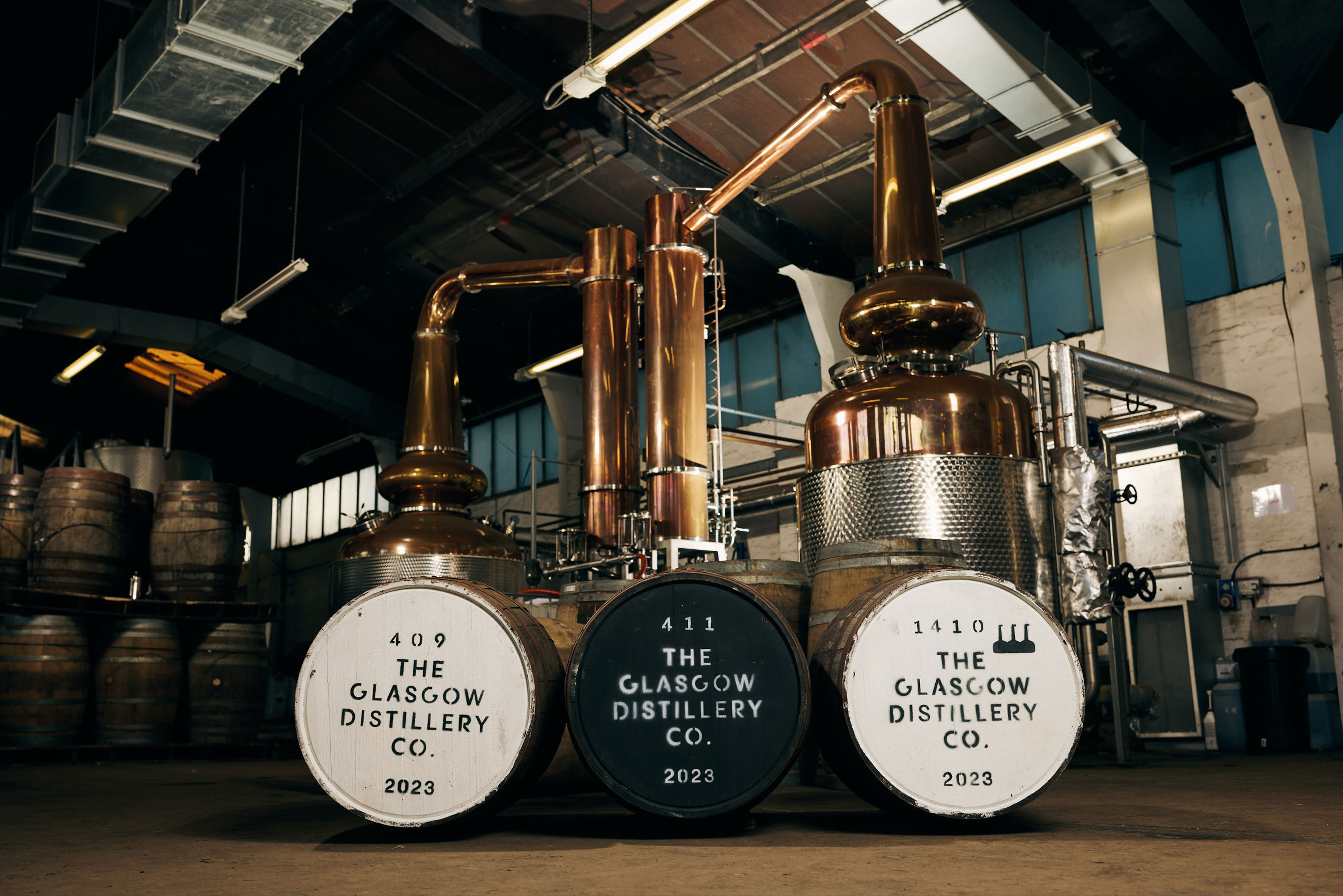 Copper stills and three large whisky barrels labeled “The Glasgow Distillery Co. 2023” are inside an industrial distillery with wooden casks stacked in the background and sunlight streaming from windows above.