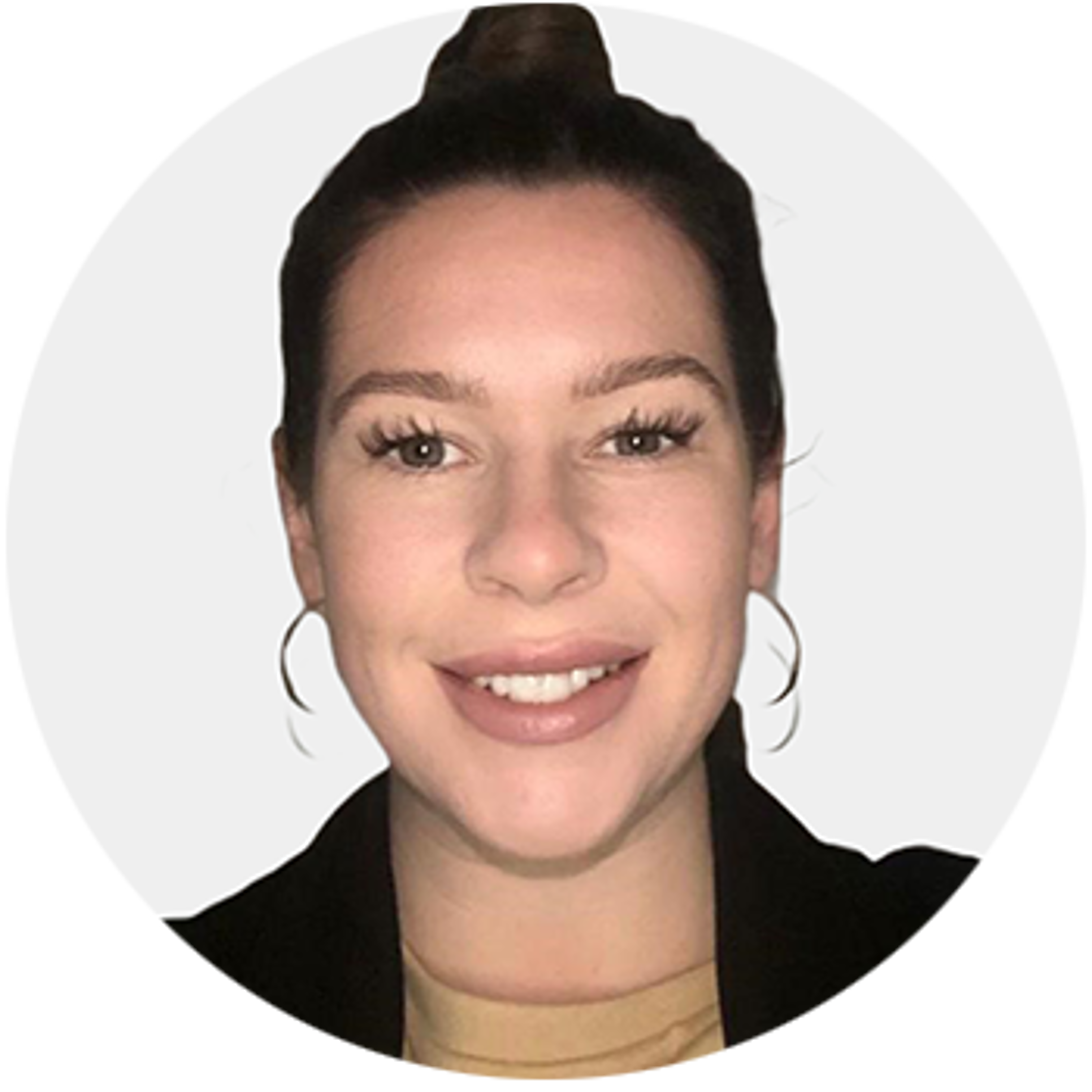 A woman with light skin and brown hair tied in a bun, wearing hoop earrings, a beige top, and a black jacket, smiling at the camera against a plain light background.