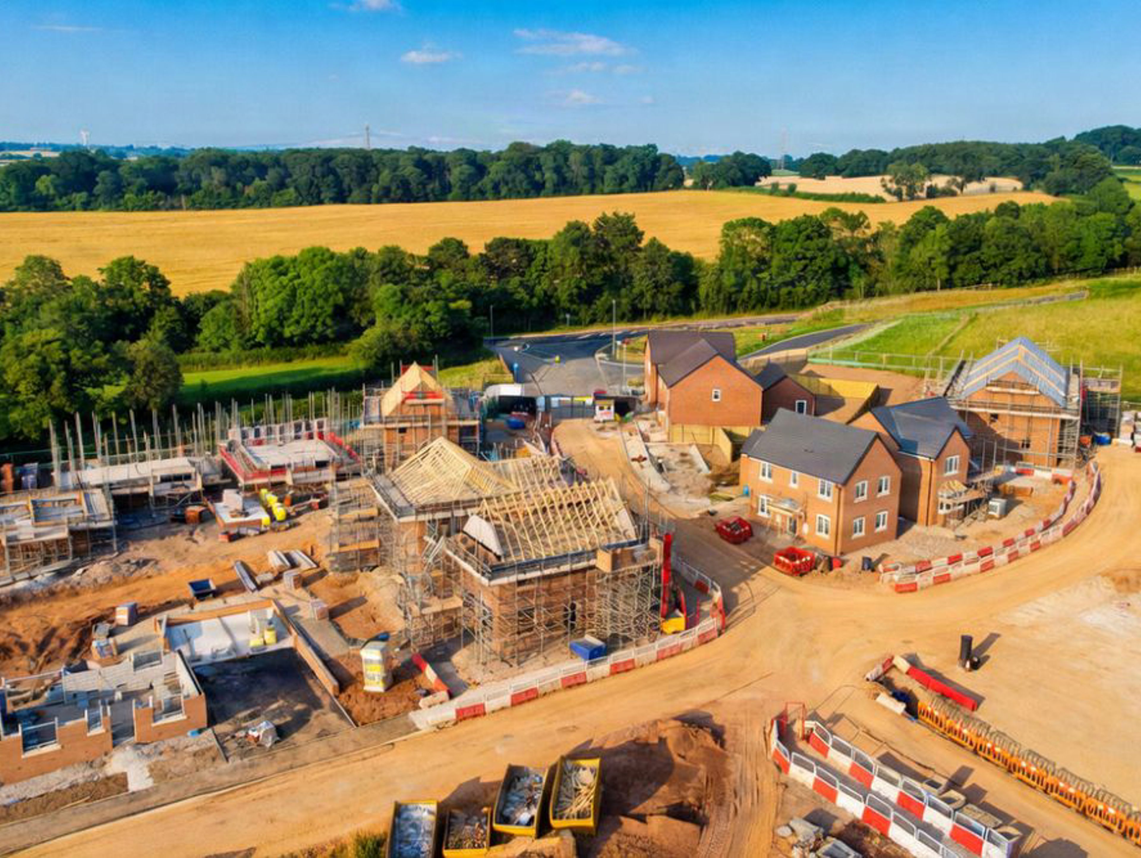 Aerial view of a residential construction site featuring several houses in various stages of completion, surrounded by fields and trees.