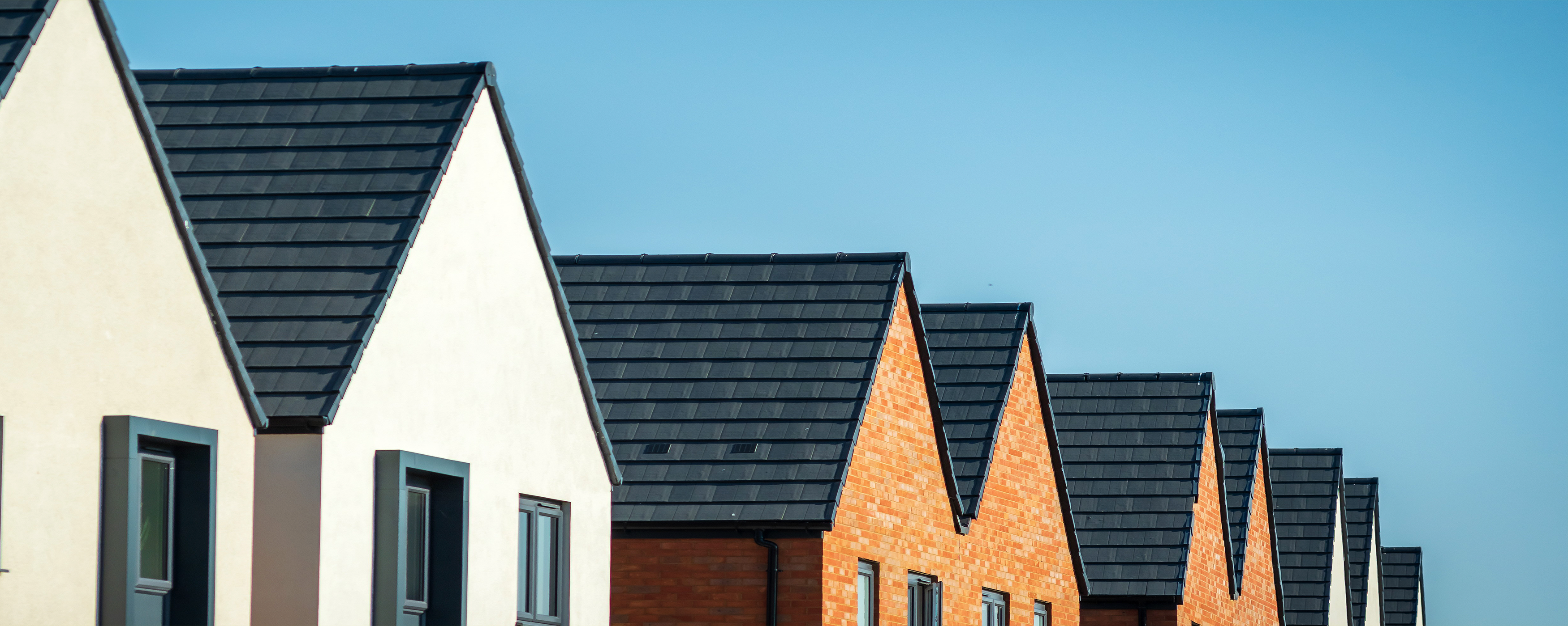 Row of modern houses with triangular, dark gray roofs and a mix of white and brick exteriors, set against a clear blue sky.