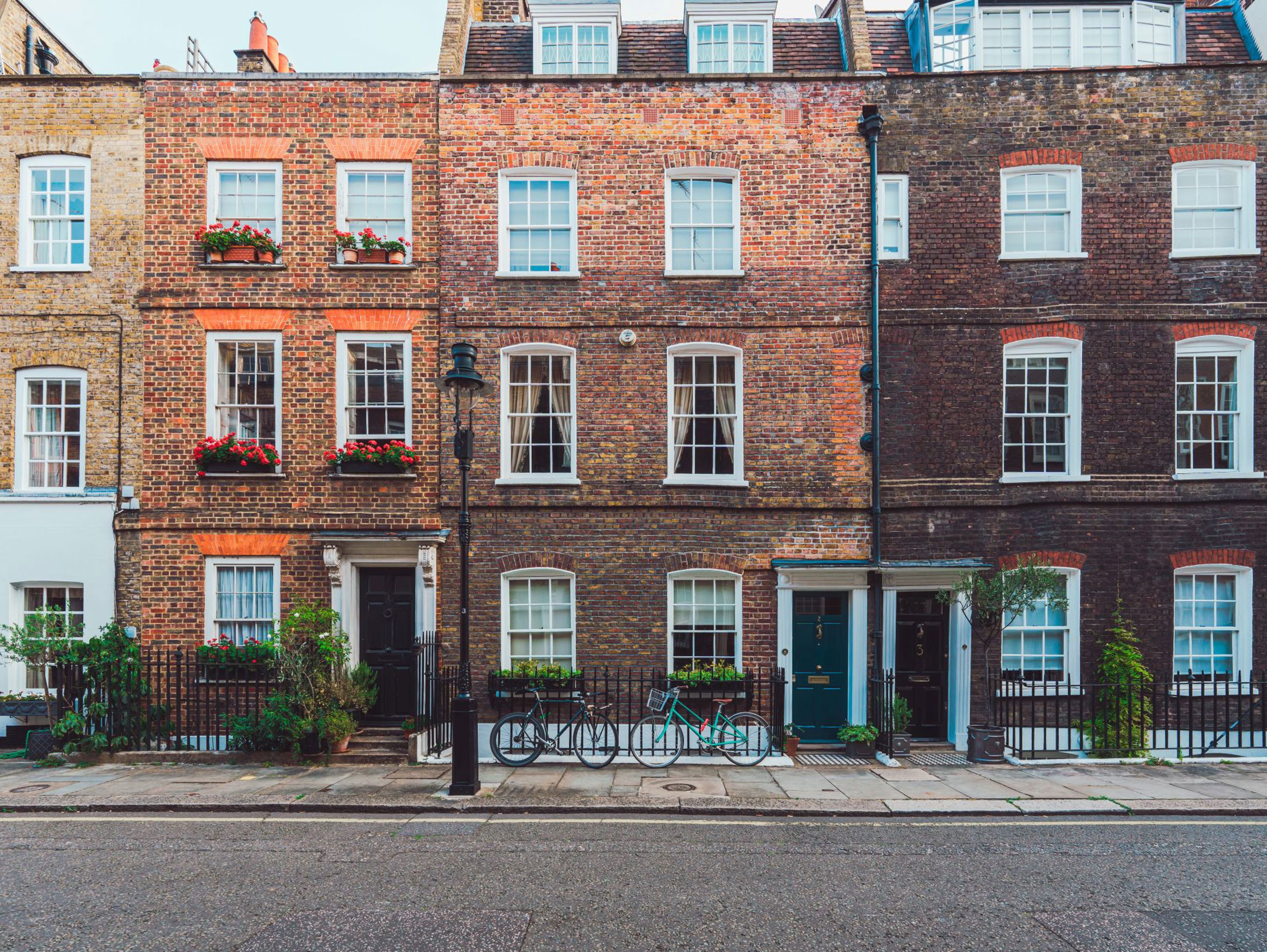 A row of colorful brick townhouses with white-framed windows, some with flower boxes, plants near the doors, and bicycles parked along a black iron fence on a quiet street.