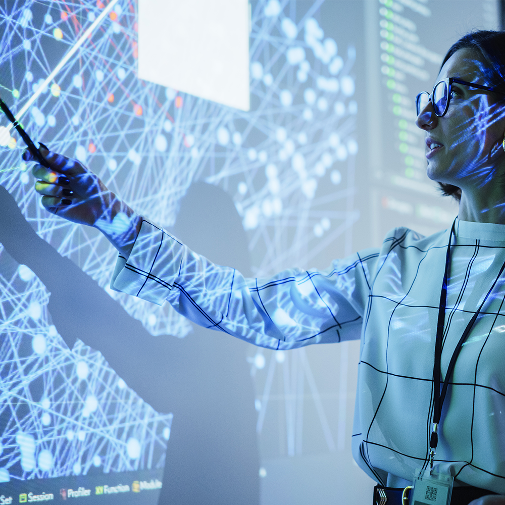 A person wearing glasses points at a large screen displaying a complex network of blue digital nodes and connections, with light patterns projected onto their clothing.