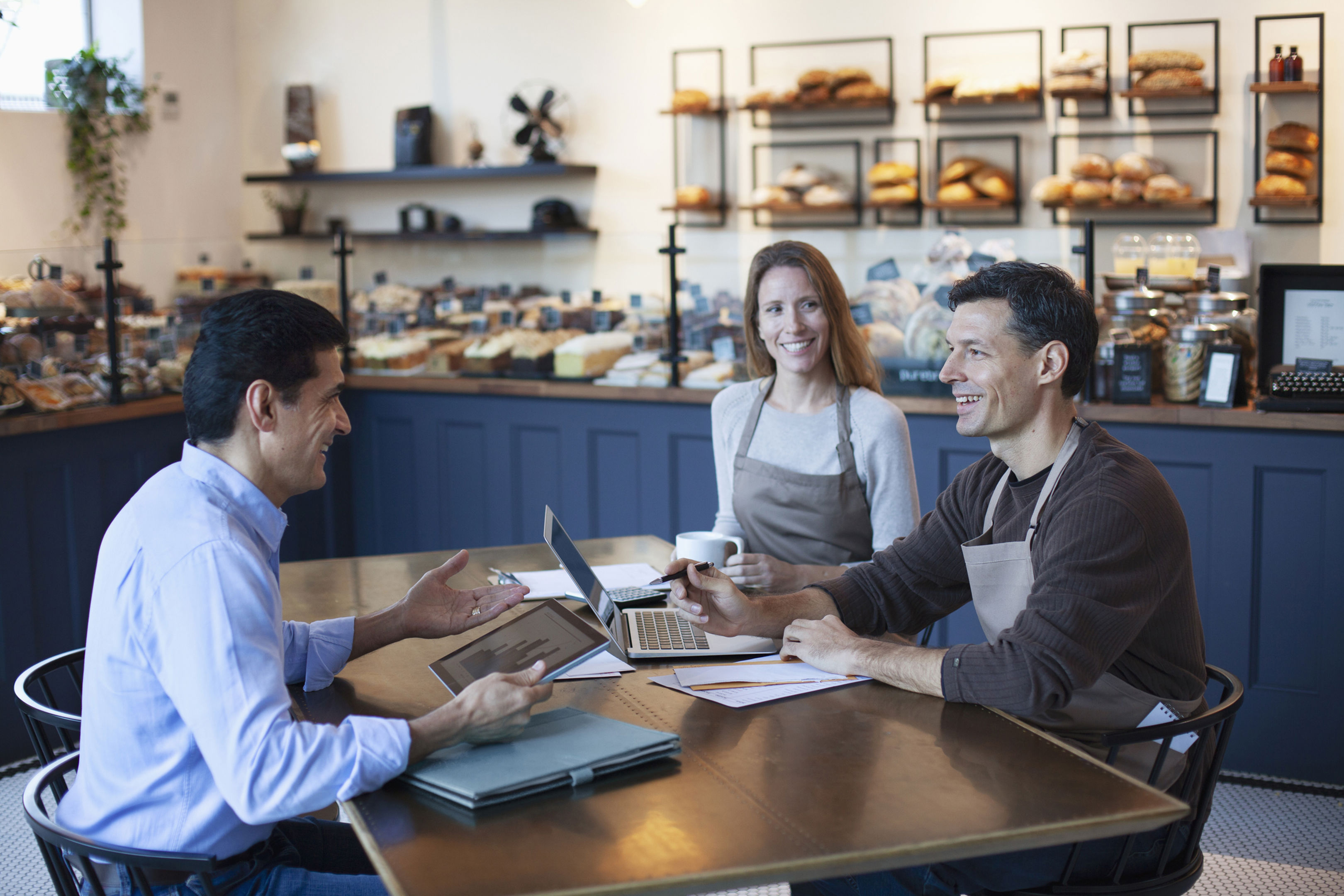 Three people sit at a table in a bakery, smiling and talking. Two wear aprons, one holds a coffee cup, and the other uses a laptop. Shelves filled with bread are visible in the background.