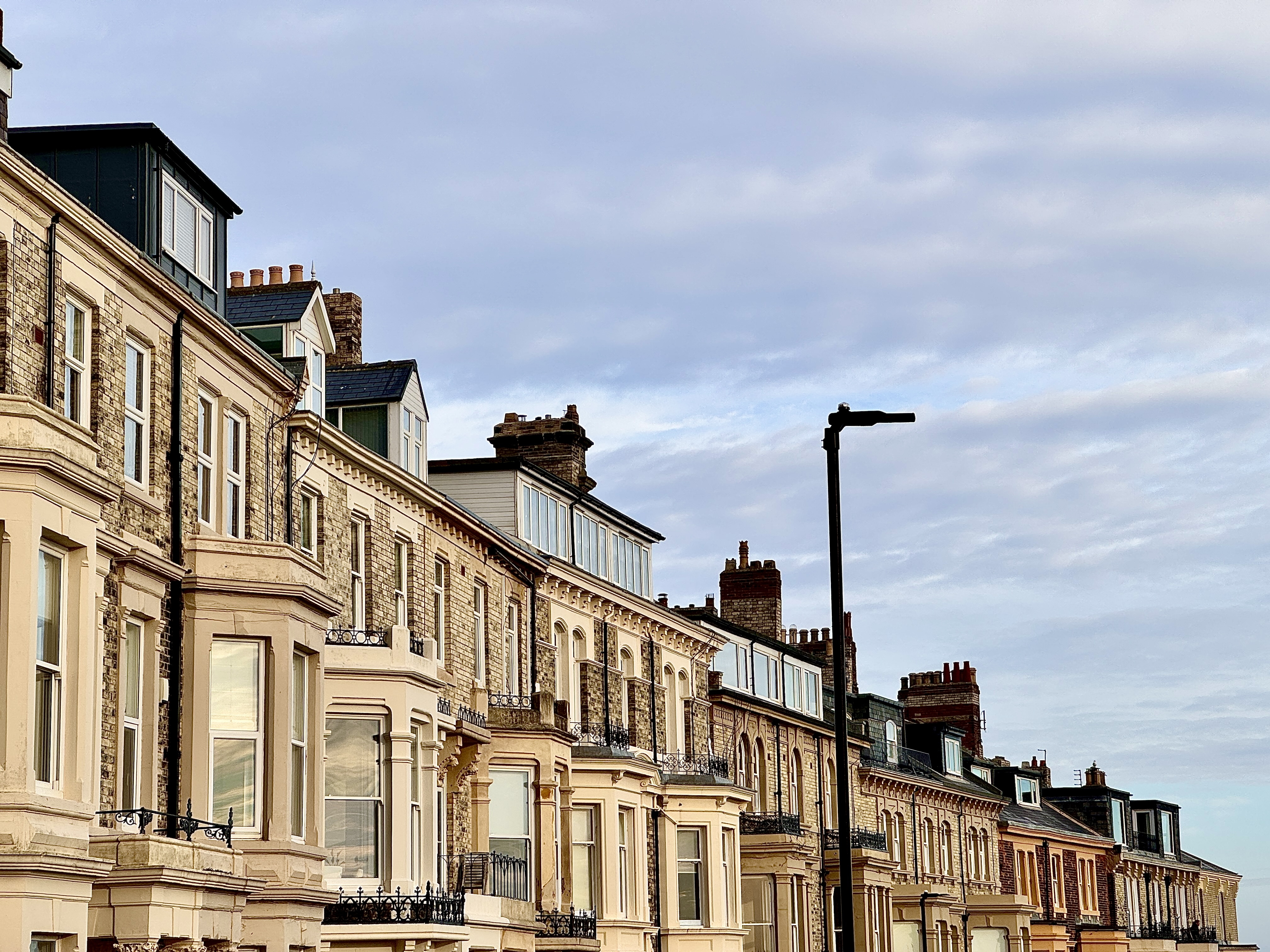 Row of Victorian-style terraced houses with brick and cream facades, large windows, and chimneys, under a partly cloudy sky. A streetlamp is visible in the foreground.