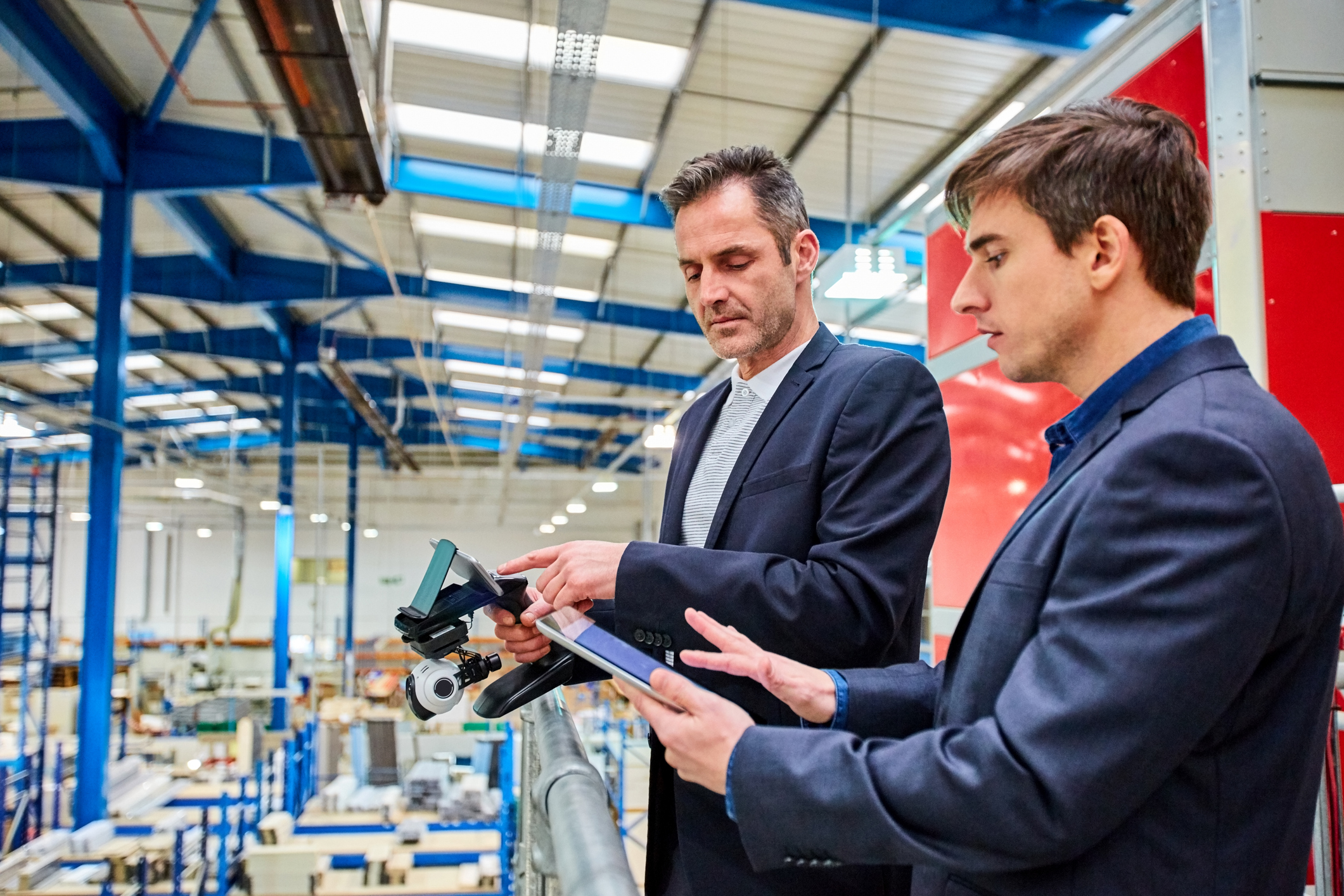 Two men in business attire stand on a balcony in a large, modern factory, looking at a tablet and drone controller, overseeing the manufacturing floor filled with equipment and machinery.