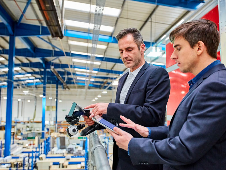 Two men in business attire stand on a balcony in a large, modern factory, looking at a tablet and drone controller, overseeing the manufacturing floor filled with equipment and machinery.