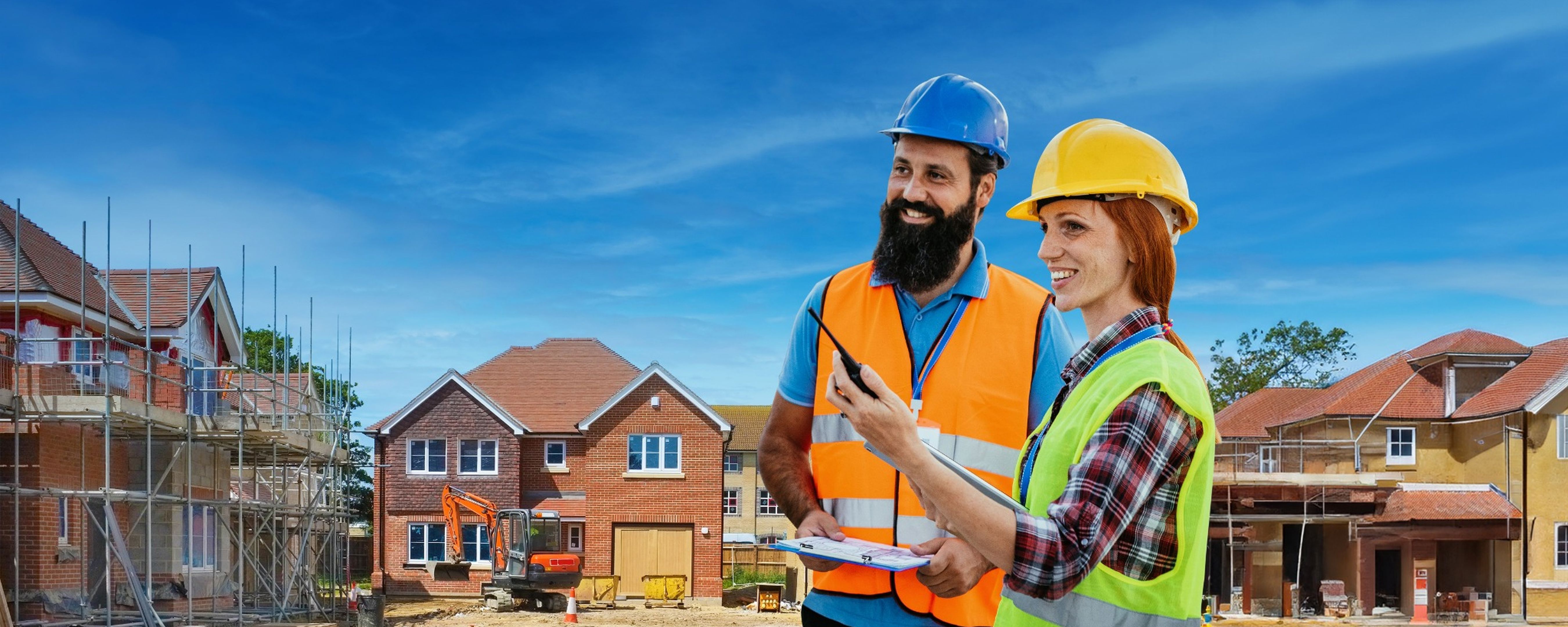 Two construction workers wearing hard hats and reflective vests smile and talk while holding a clipboard and walkie-talkie at a residential construction site with houses and scaffolding in the background.