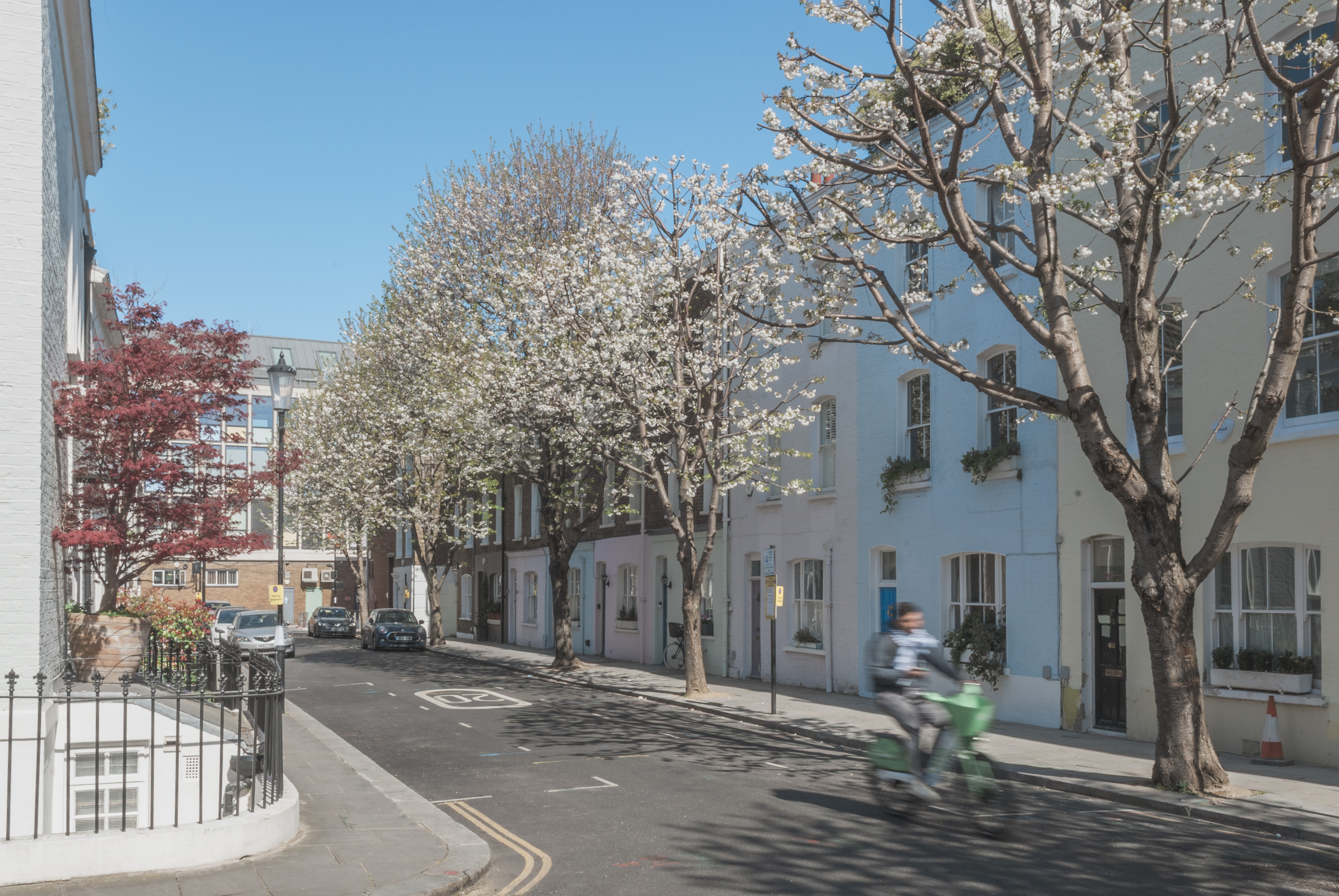 A quiet, sunny street lined with pastel-colored houses and blossoming trees. A person on a green scooter rides past, and a few parked cars are visible under the blue sky.