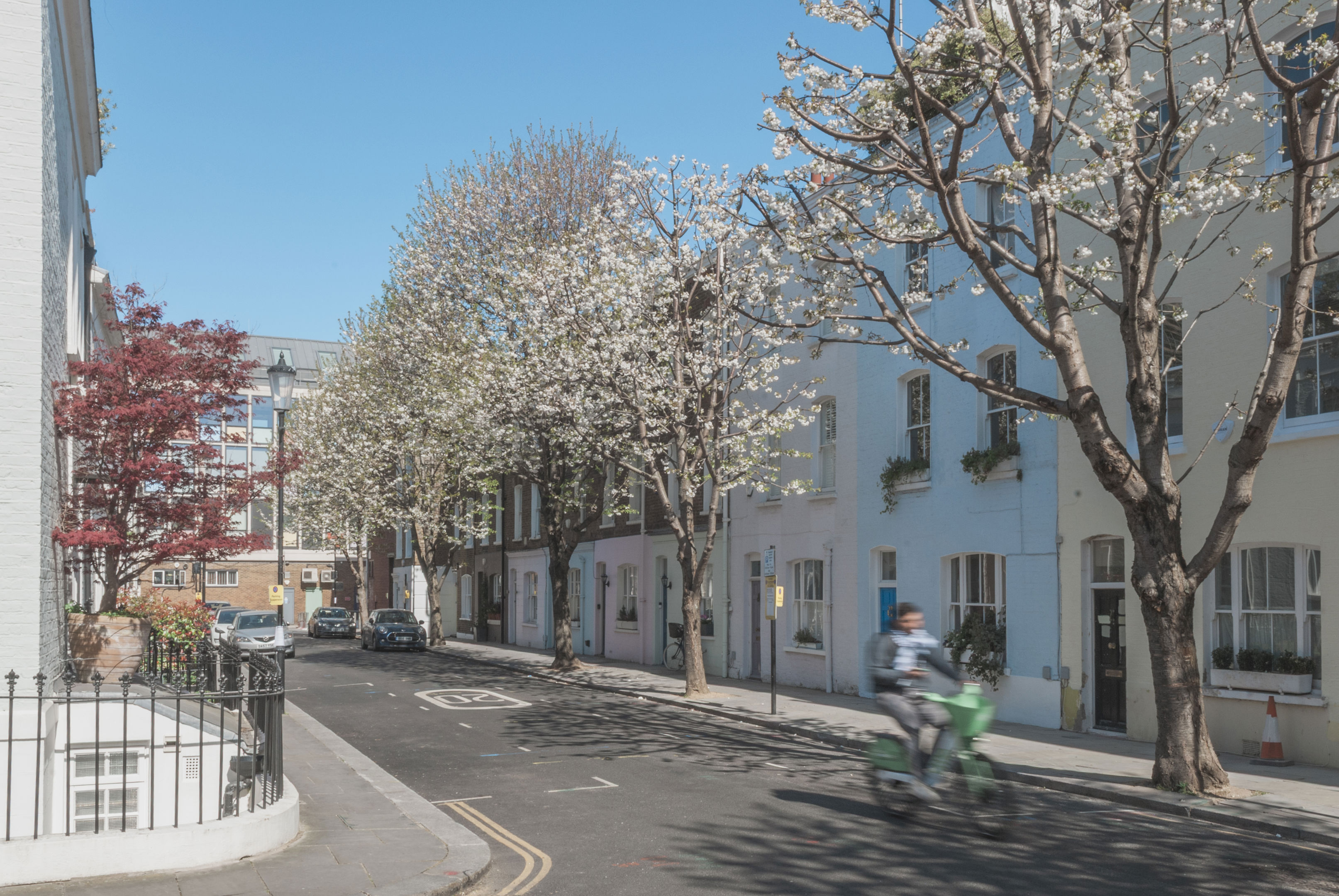 A quiet, sunny street lined with pastel-colored houses and blossoming trees. A person on a green scooter rides past, and a few parked cars are visible under the blue sky.