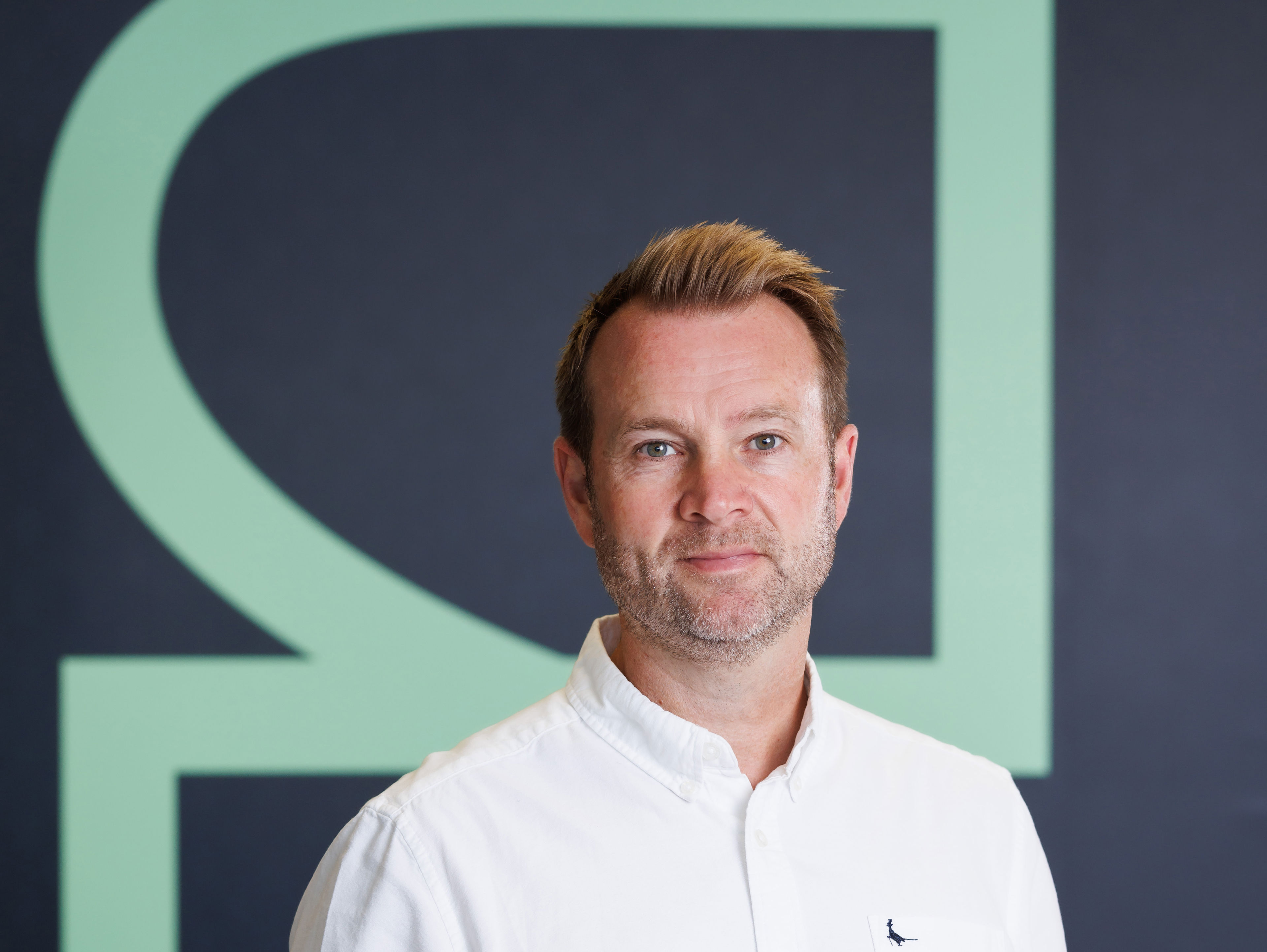 A man wearing a white button-up shirt stands in front of a dark wall with a large, abstract green geometric design. He has short light brown hair and a neutral expression.