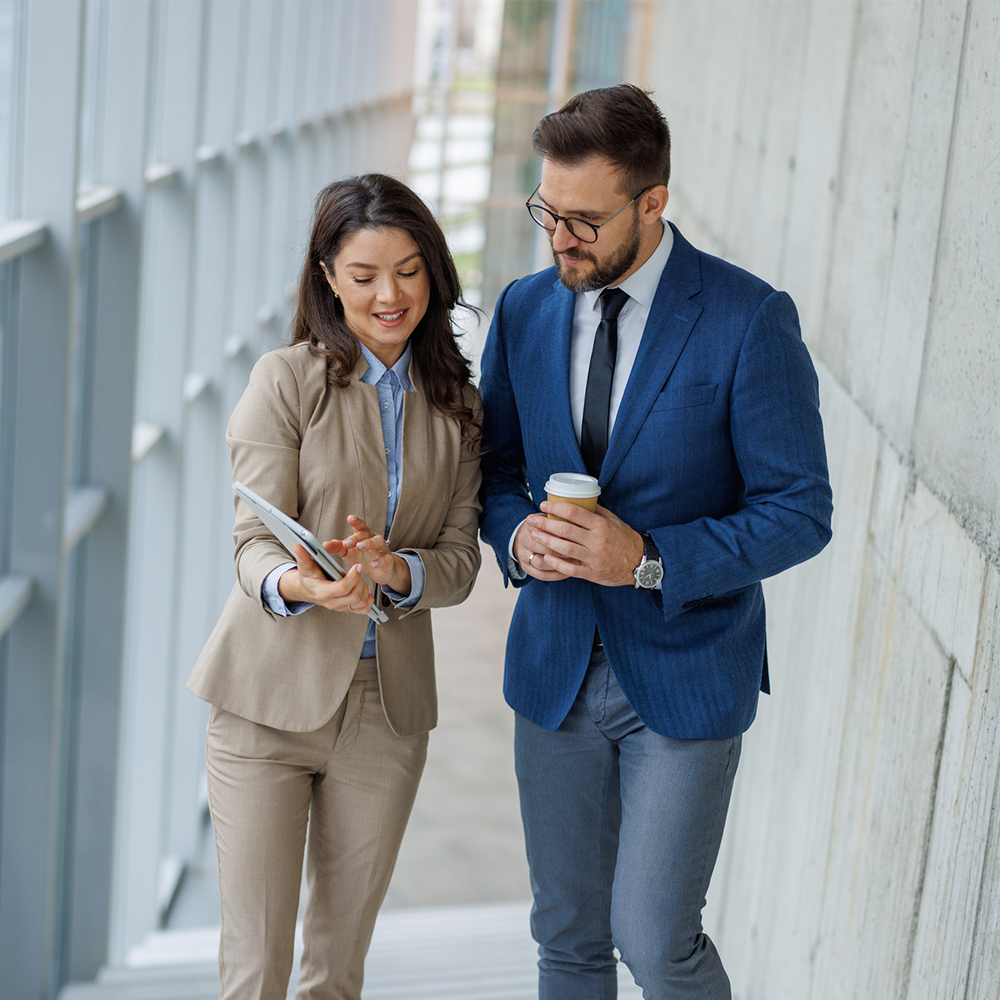 A woman in a beige suit showing a tablet to a man in a blue blazer and tie holding a coffee cup while walking through an office hallway.