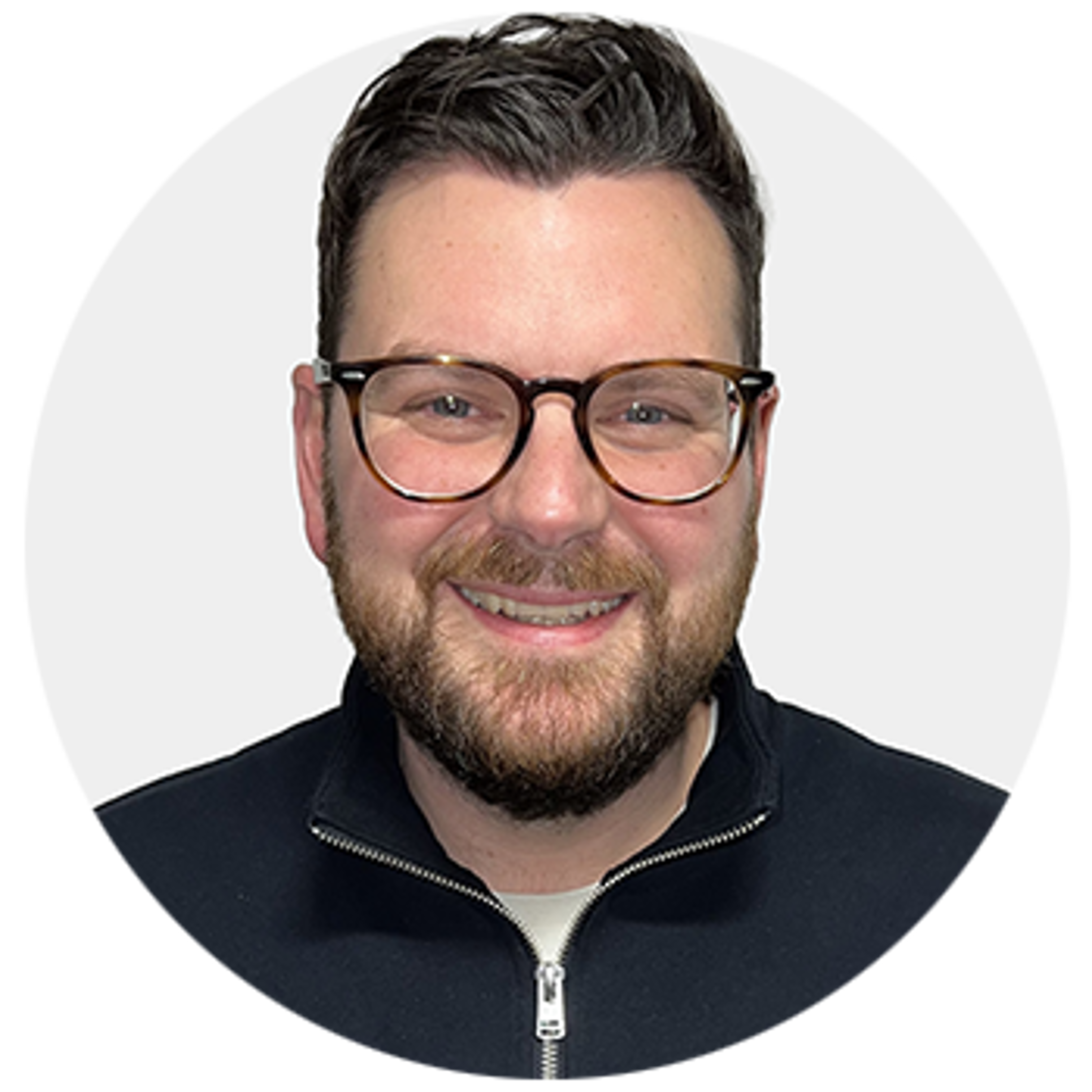 Smiling man with short dark hair, beard, and glasses, wearing a black zip-up jacket over a light shirt, posed against a plain light background.