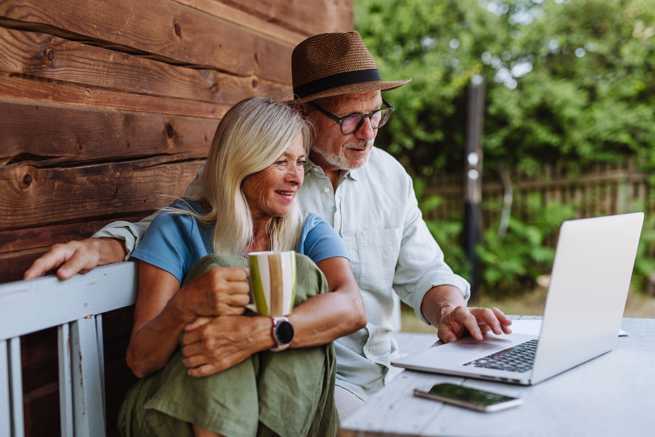 Elderly couple seated on a bench outside a wooden cabin, looking at a laptop. The woman holds a striped mug, wearing a blue shirt and green pants. The man is in a white shirt and straw hat, pointing at the screen. Greenery is visible in the background.