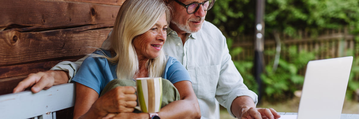 Elderly couple seated on a bench outside a wooden cabin, looking at a laptop. The woman holds a striped mug, wearing a blue shirt and green pants. The man is in a white shirt and straw hat, pointing at the screen. Greenery is visible in the background.