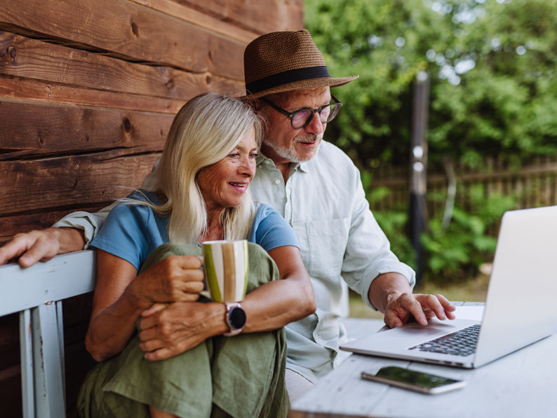 Elderly couple seated on a bench outside a wooden cabin, looking at a laptop. The woman holds a striped mug, wearing a blue shirt and green pants. The man is in a white shirt and straw hat, pointing at the screen. Greenery is visible in the background.