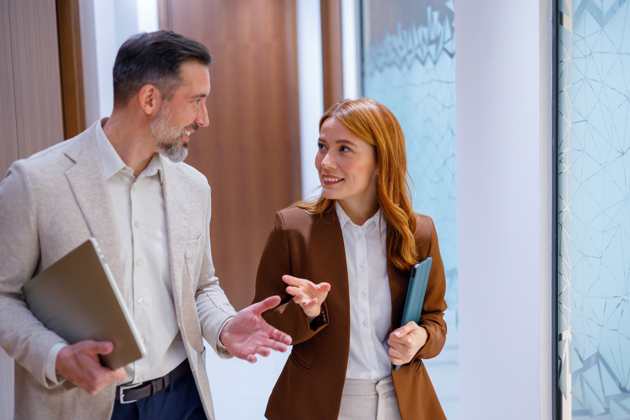 A man and a woman in business attire walking and talking in an office hallway while carrying tablets.