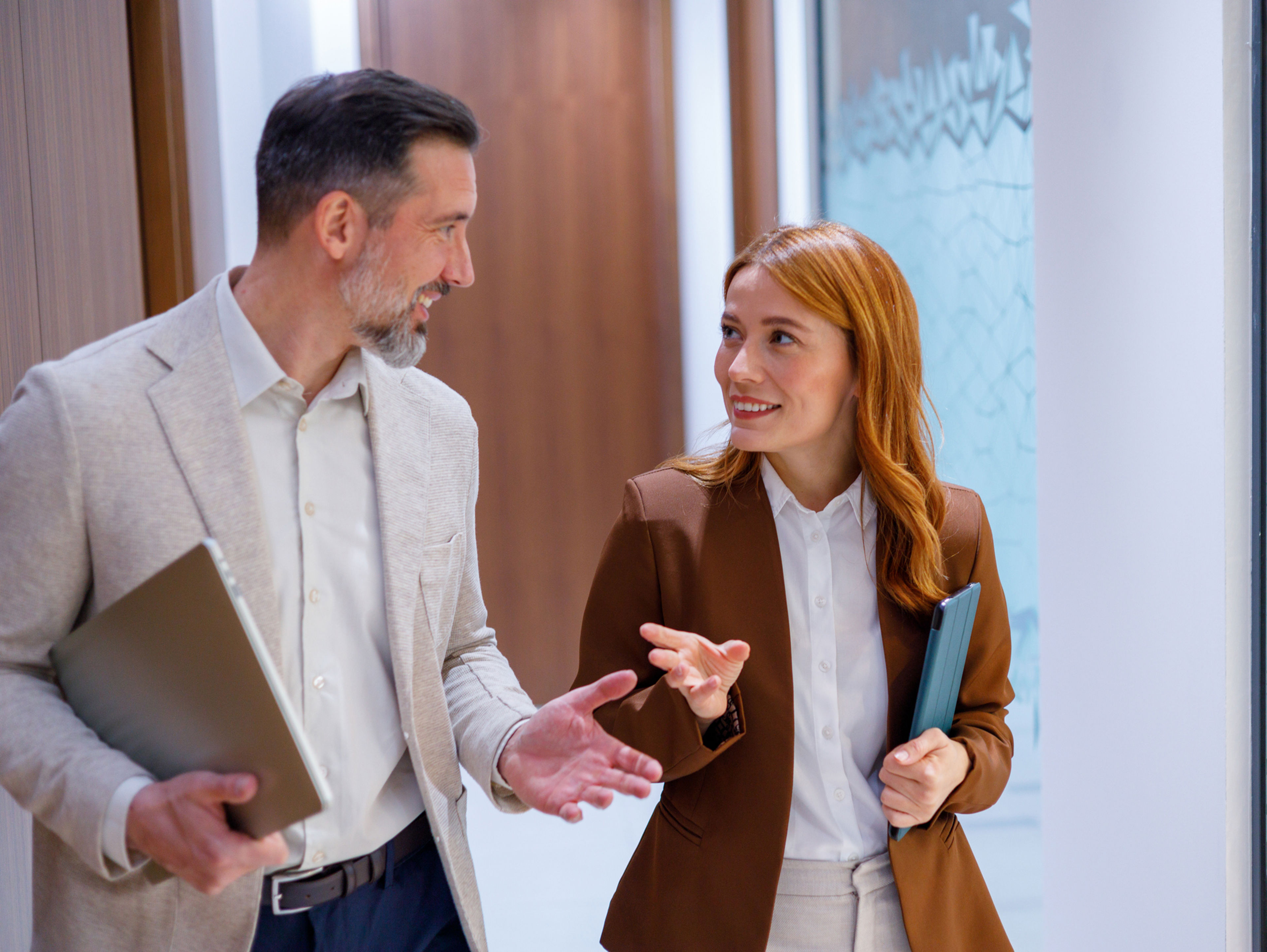 A man and a woman in business attire walking and talking in an office hallway while carrying tablets.
