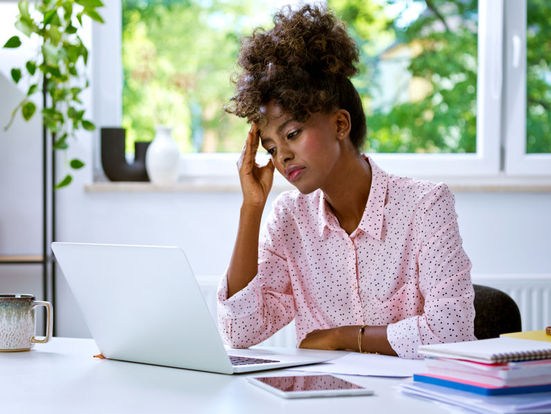 A woman sits at a desk, looking stressed while holding her forehead and staring at a laptop screen. Notebooks, a tablet, and plants are on the desk, with large windows showing greenery in the background.