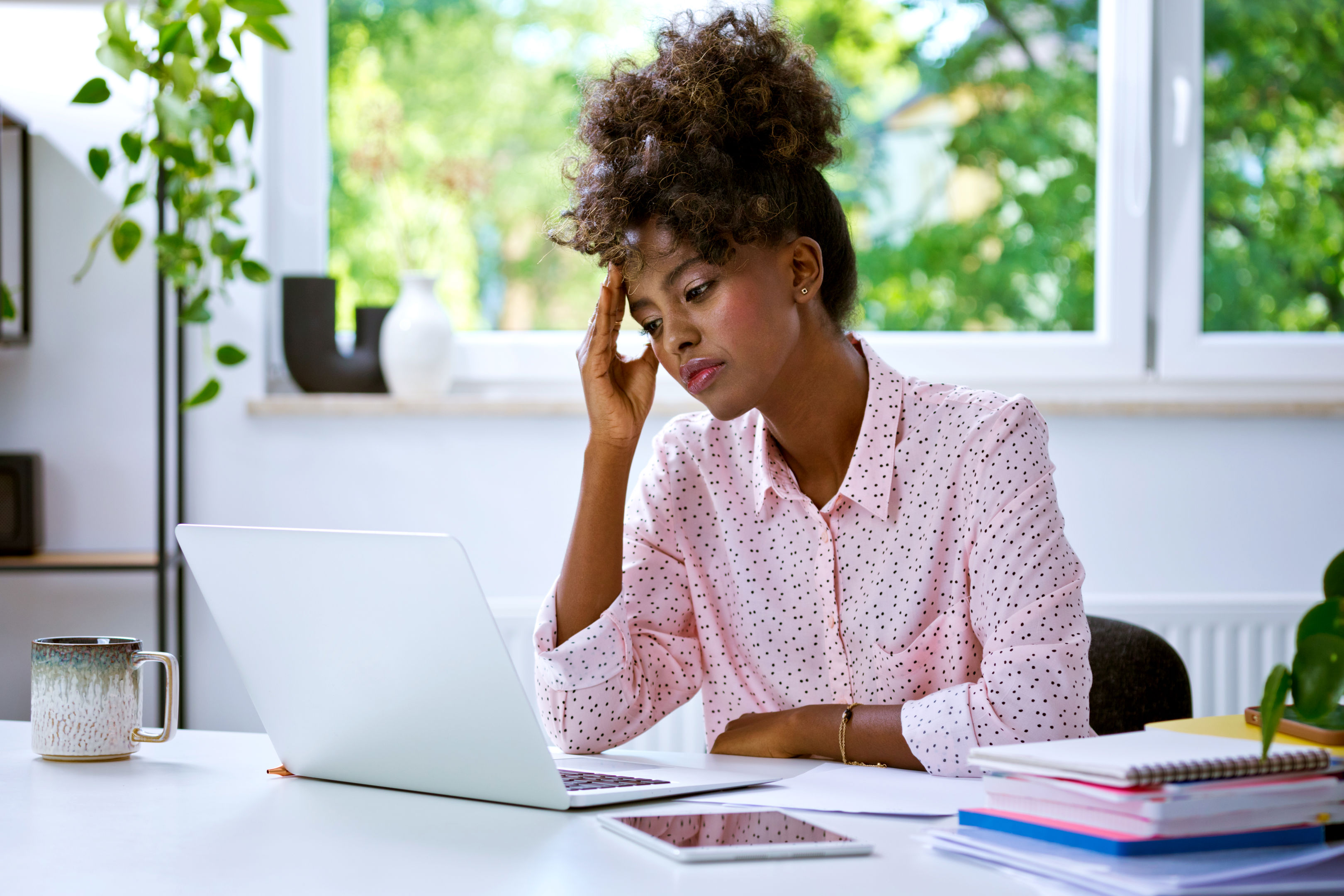 A woman sits at a desk, looking stressed while holding her forehead and staring at a laptop screen. Notebooks, a tablet, and plants are on the desk, with large windows showing greenery in the background.