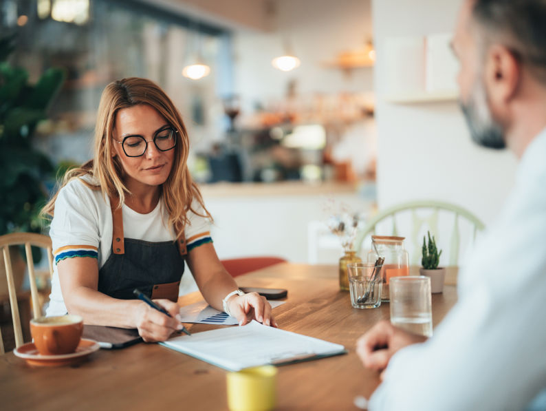 A woman wearing glasses and an apron sits at a table in a bright café, writing on a clipboard, while a man sits across from her. There are coffee cups, water glasses, and plants on the table.