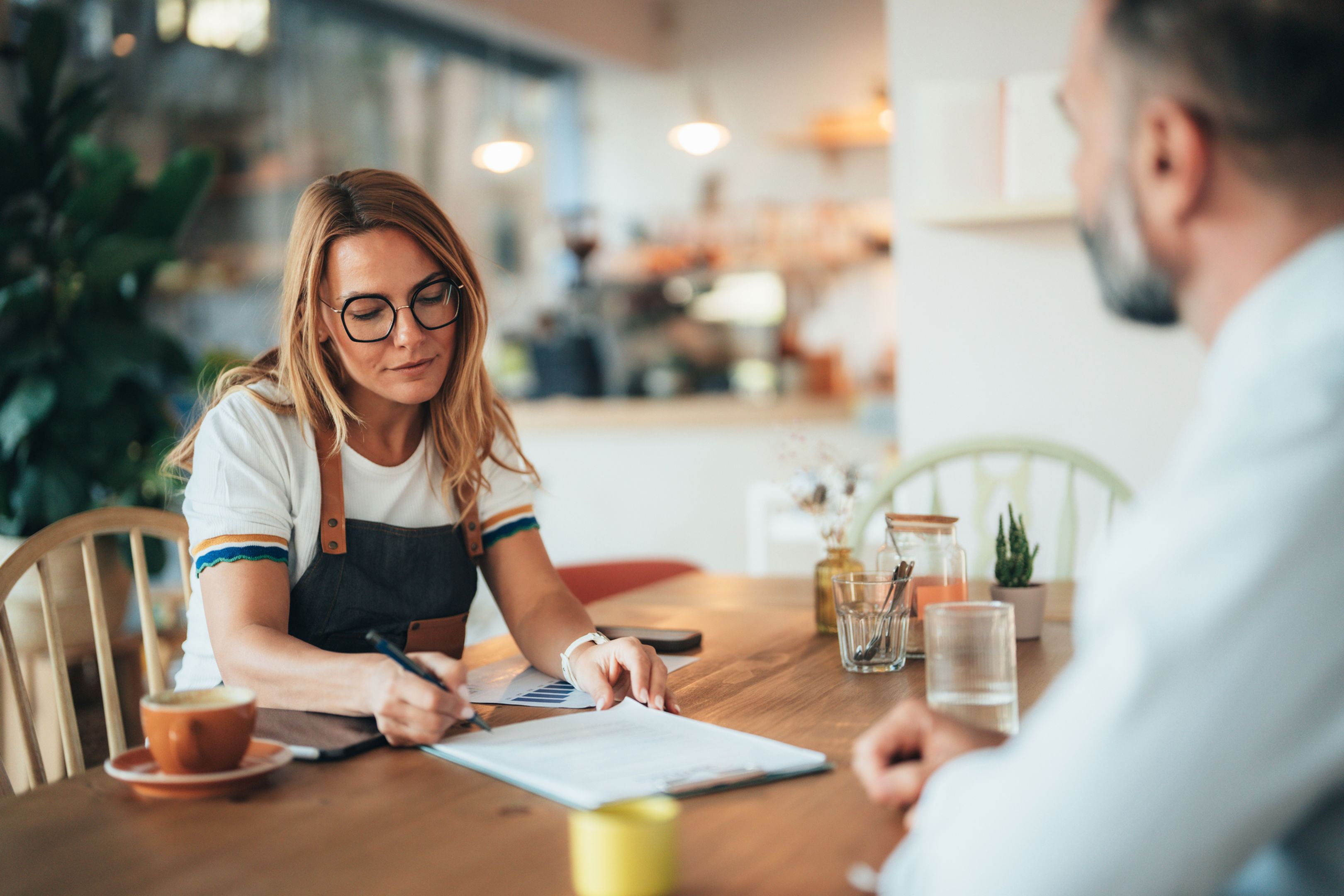 A woman wearing glasses and an apron sits at a table in a bright café, writing on a clipboard, while a man sits across from her. There are coffee cups, water glasses, and plants on the table.