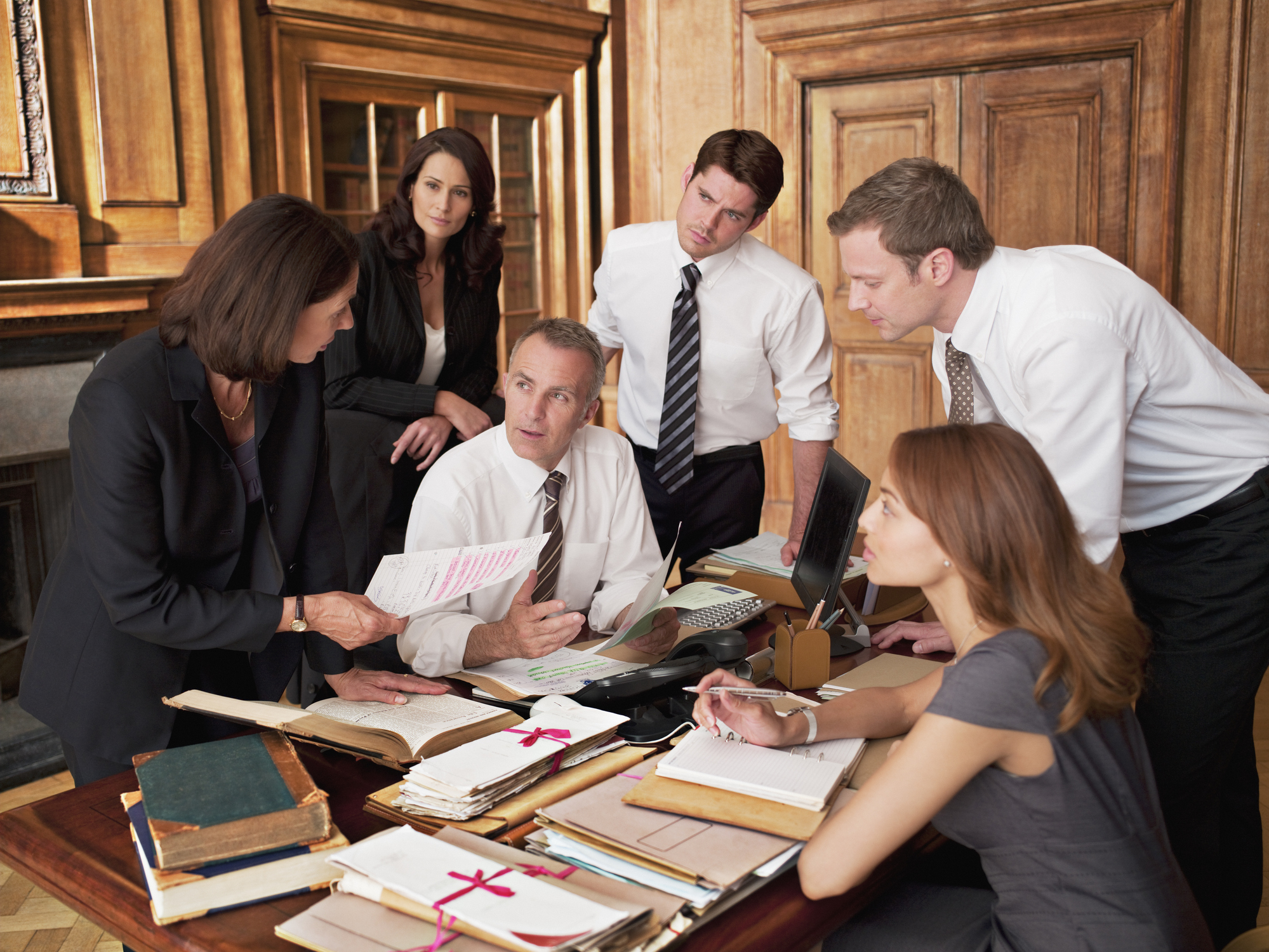 Five professionals in formal attire gather around a desk covered with documents, folders, and books, engaged in a serious discussion in a wood-paneled office, with one woman presenting papers to the group.
