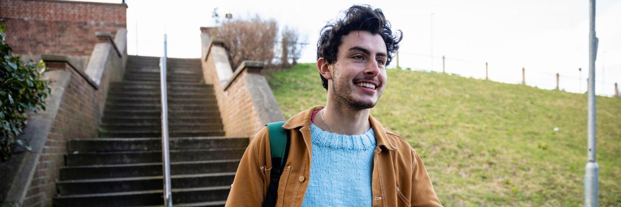 A smiling young man wearing a light blue sweater and brown jacket walks outside while holding a smartphone.