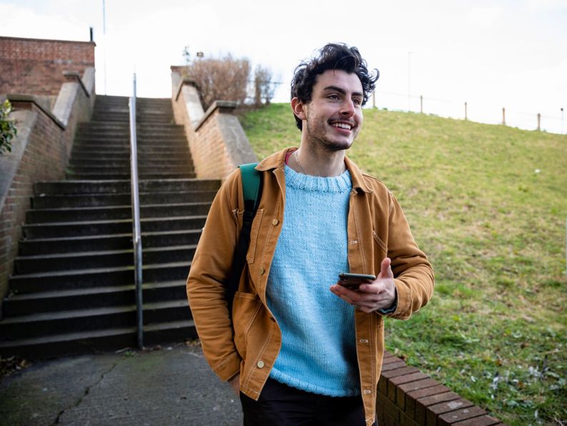 A smiling young man wearing a light blue sweater and brown jacket walks outside while holding a smartphone.