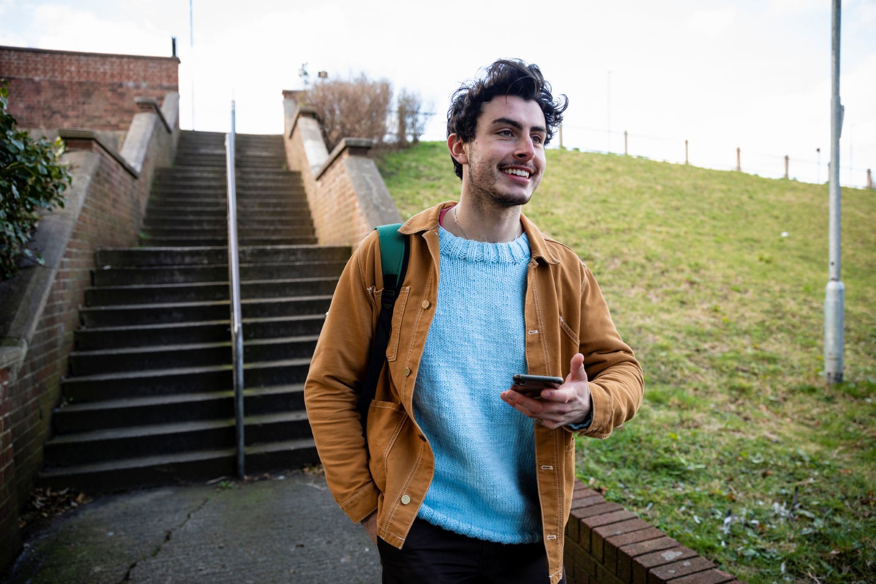 A smiling young man wearing a light blue sweater and brown jacket walks outside while holding a smartphone.