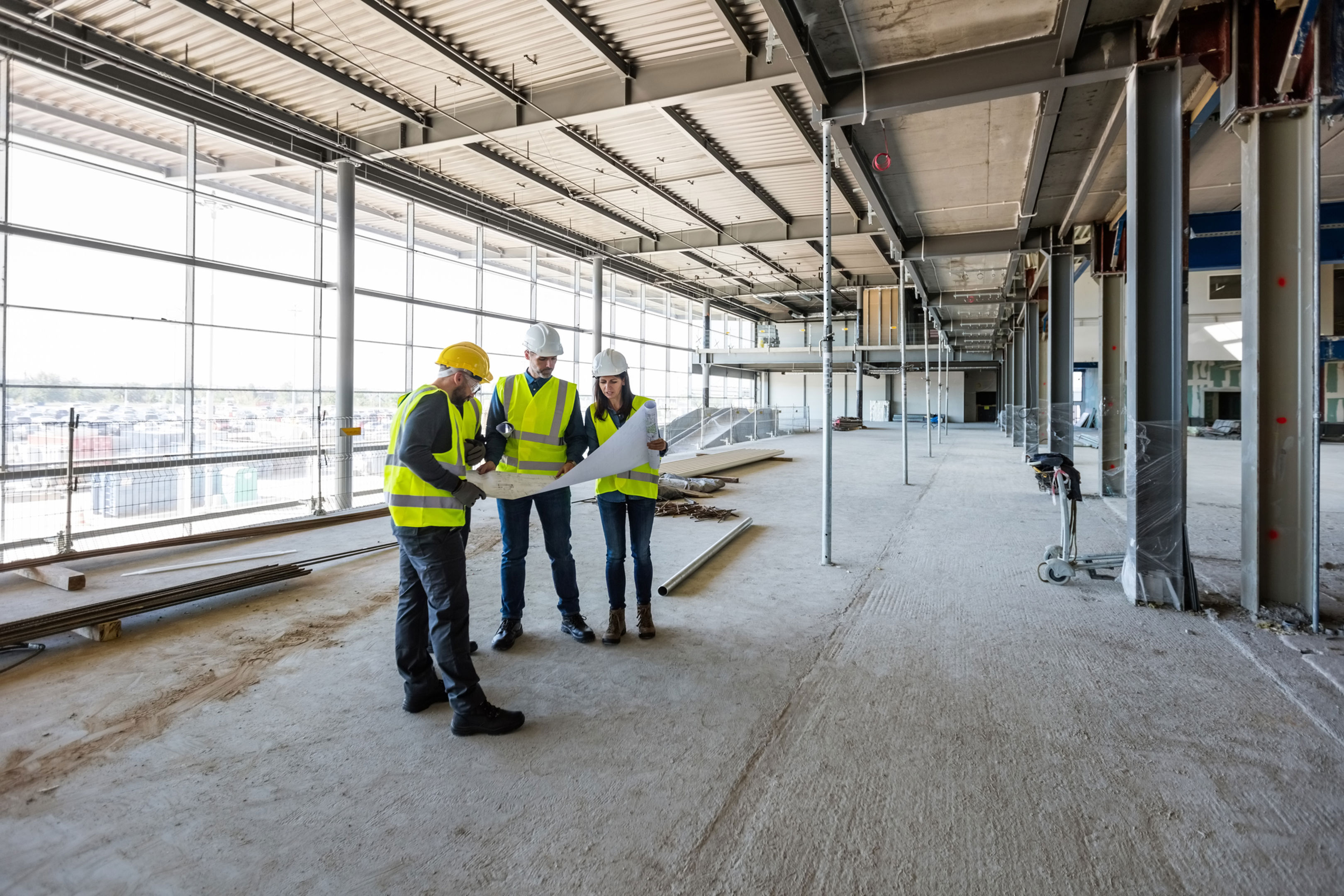 Three construction workers in safety vests and helmets review blueprints inside a large, unfinished building with exposed beams, concrete floors, and large windows letting in natural light.