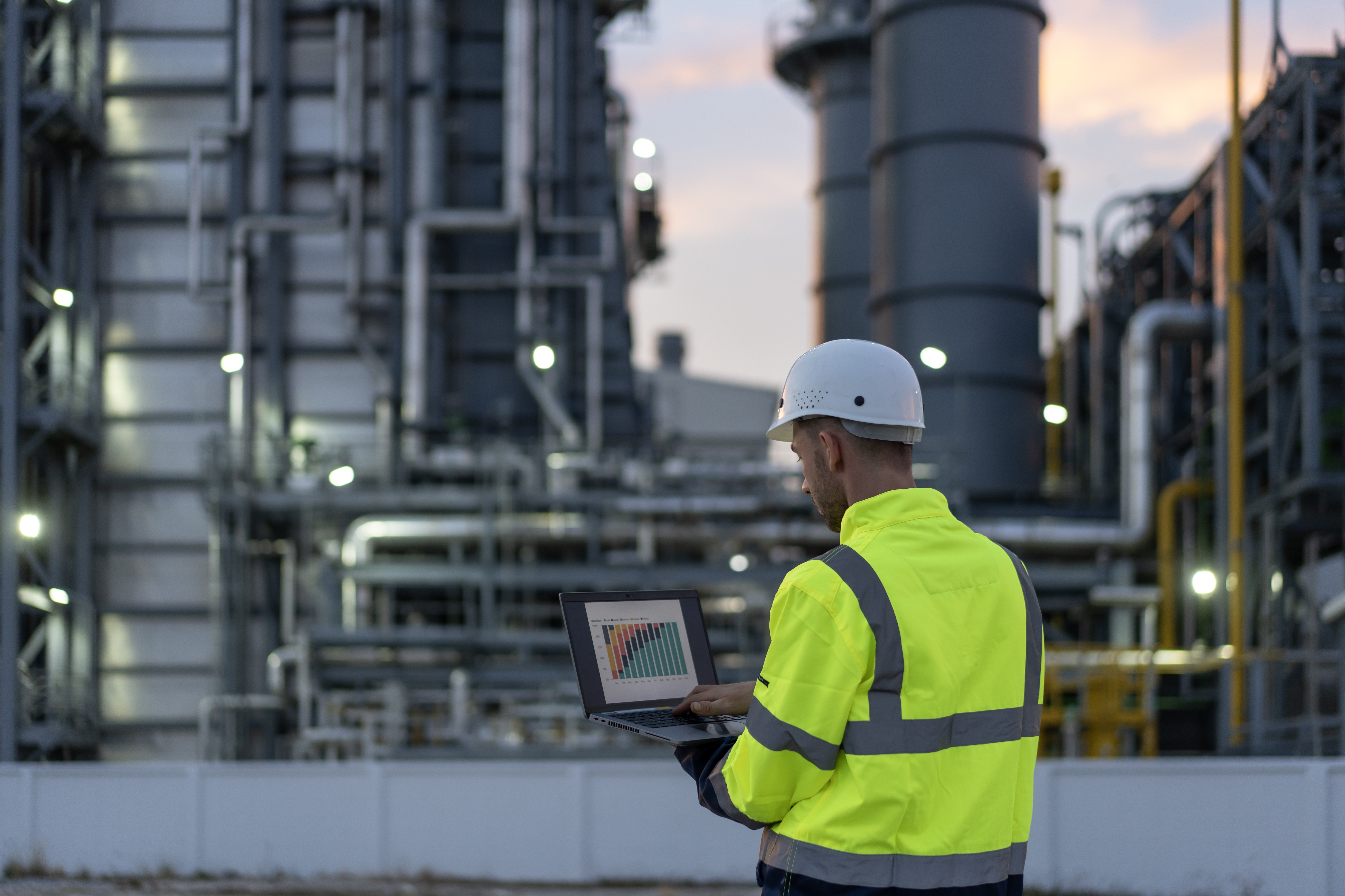 A worker in a high-visibility jacket and helmet uses a laptop with a bar graph on the screen at an industrial plant with large machinery and pipes in the background.