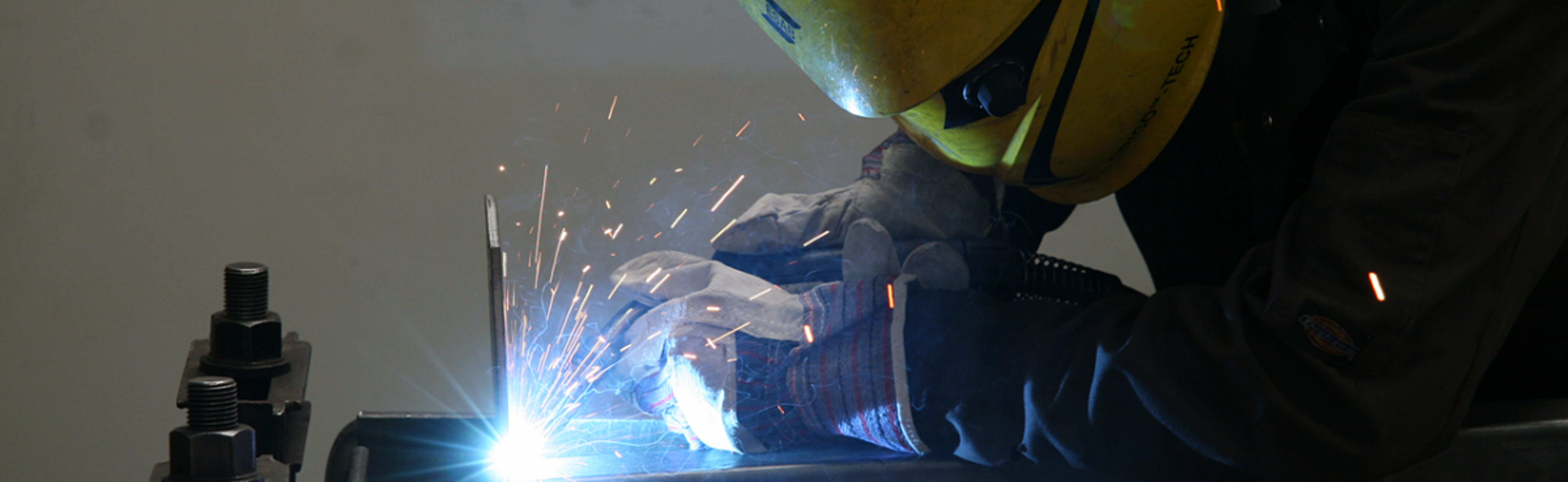 A person wearing protective gloves and a yellow welding helmet is welding metal, with bright sparks flying from the contact point.