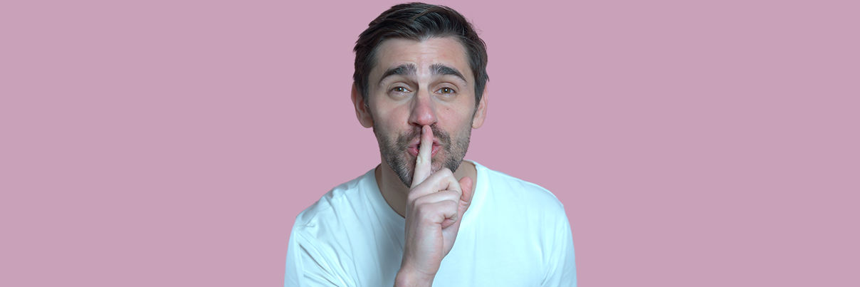 A person with dark hair and beard, wearing a white T-shirt, holds their index finger to their lips in a shushing gesture, against a solid pink background.